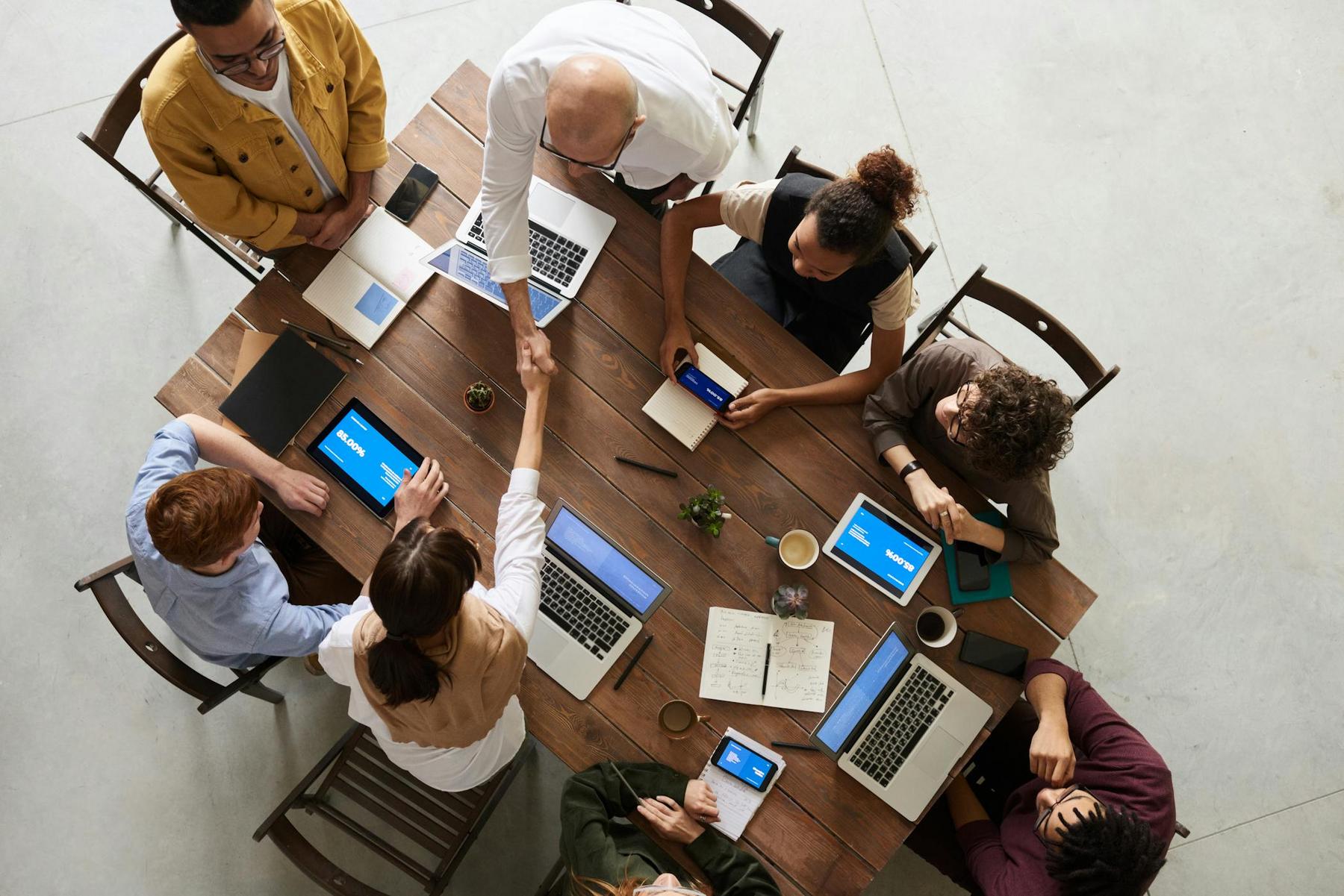 A group of people sit around a wooden table with laptops and notebooks, while two individuals in the center shake hands.