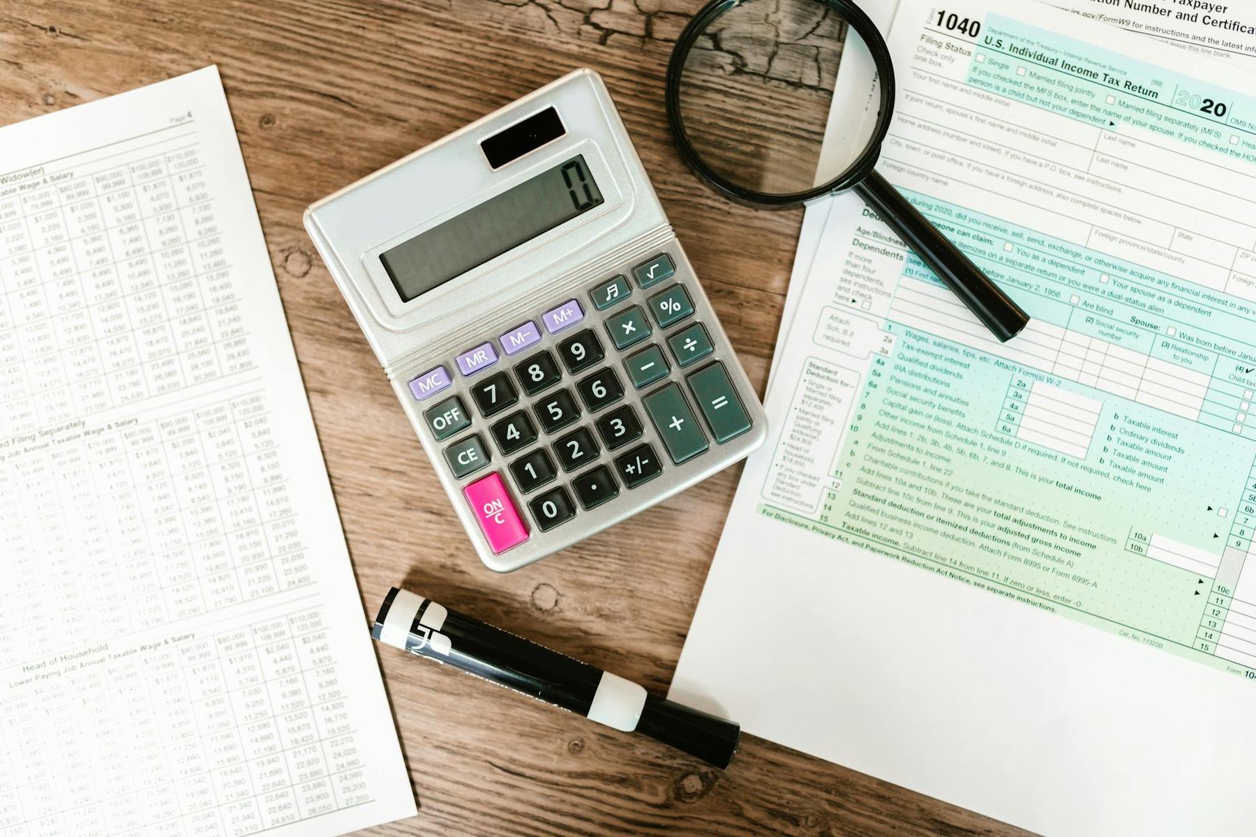 A calculator, magnifying glass, pen, tax tables, and a completed IRS 1040 tax form are arranged on a wooden desk.