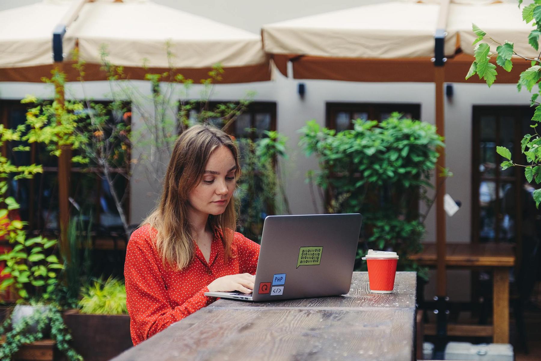 A woman in a red shirt works on a laptop at an outdoor wooden table with a red paper cup beside her, surrounded by plants and umbrellas.