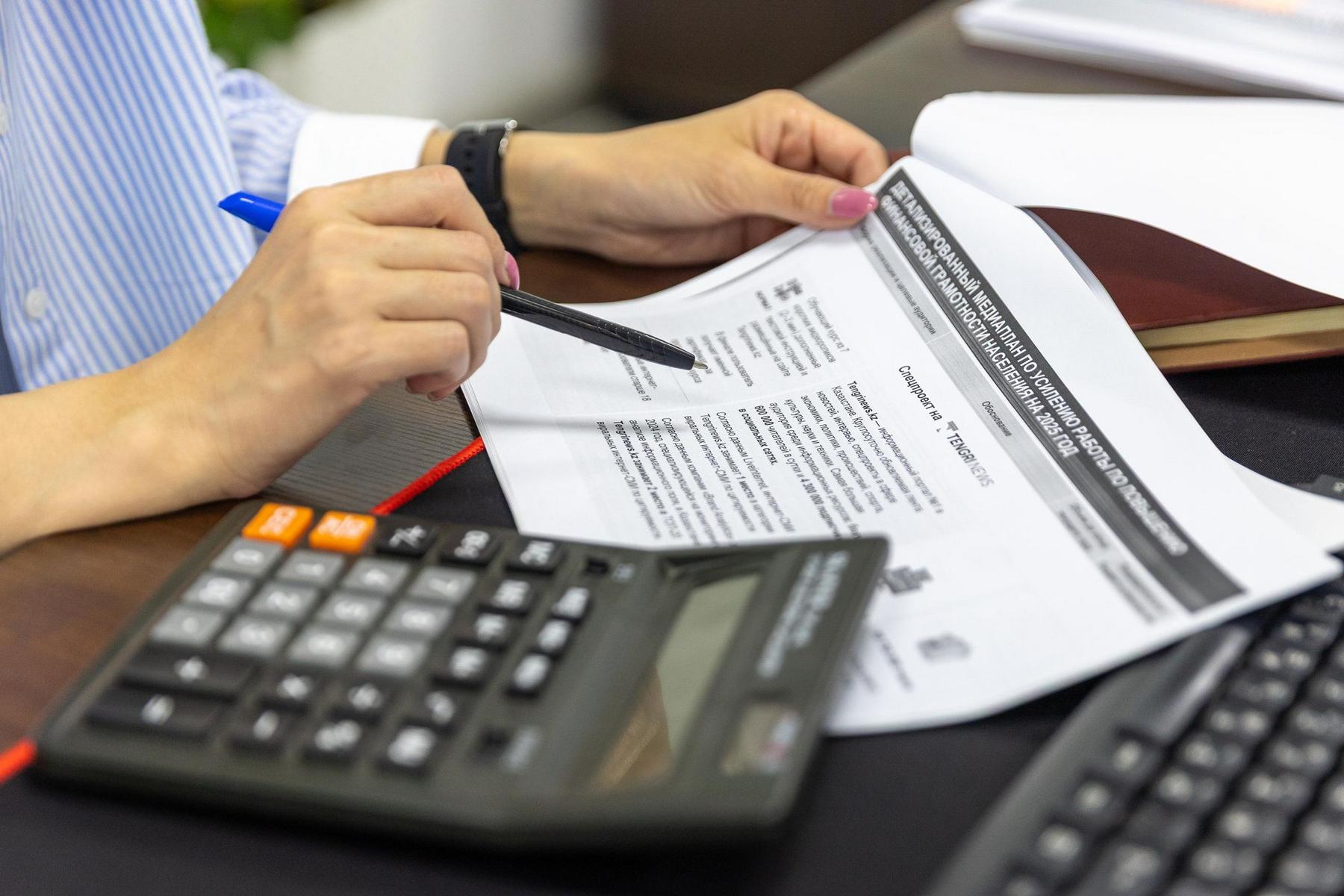 A person holds a pen while reviewing a printed document, with a calculator and a notebook on the desk.