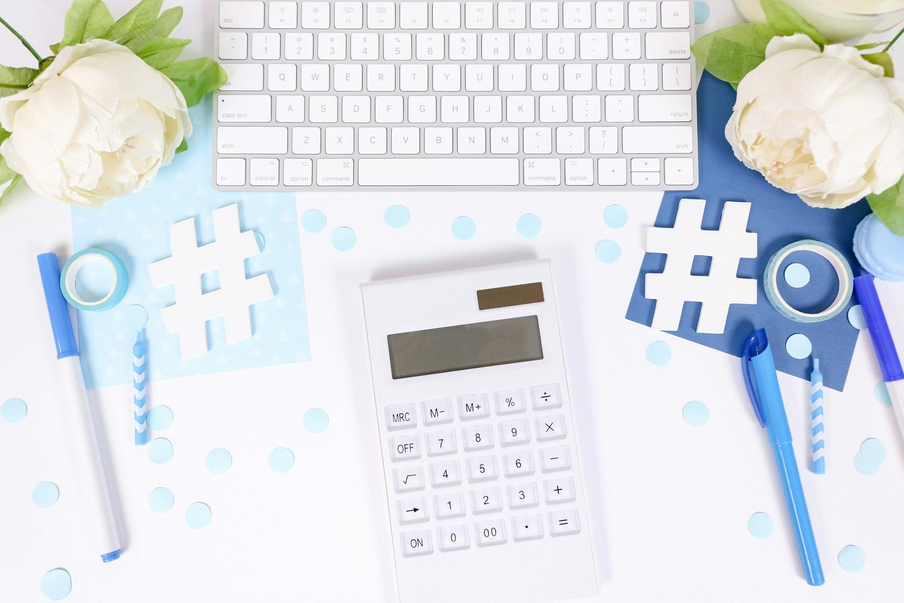 A white calculator rests on a desk with a keyboard, paper hashtag cutouts, pens, washi tape, and white flowers.