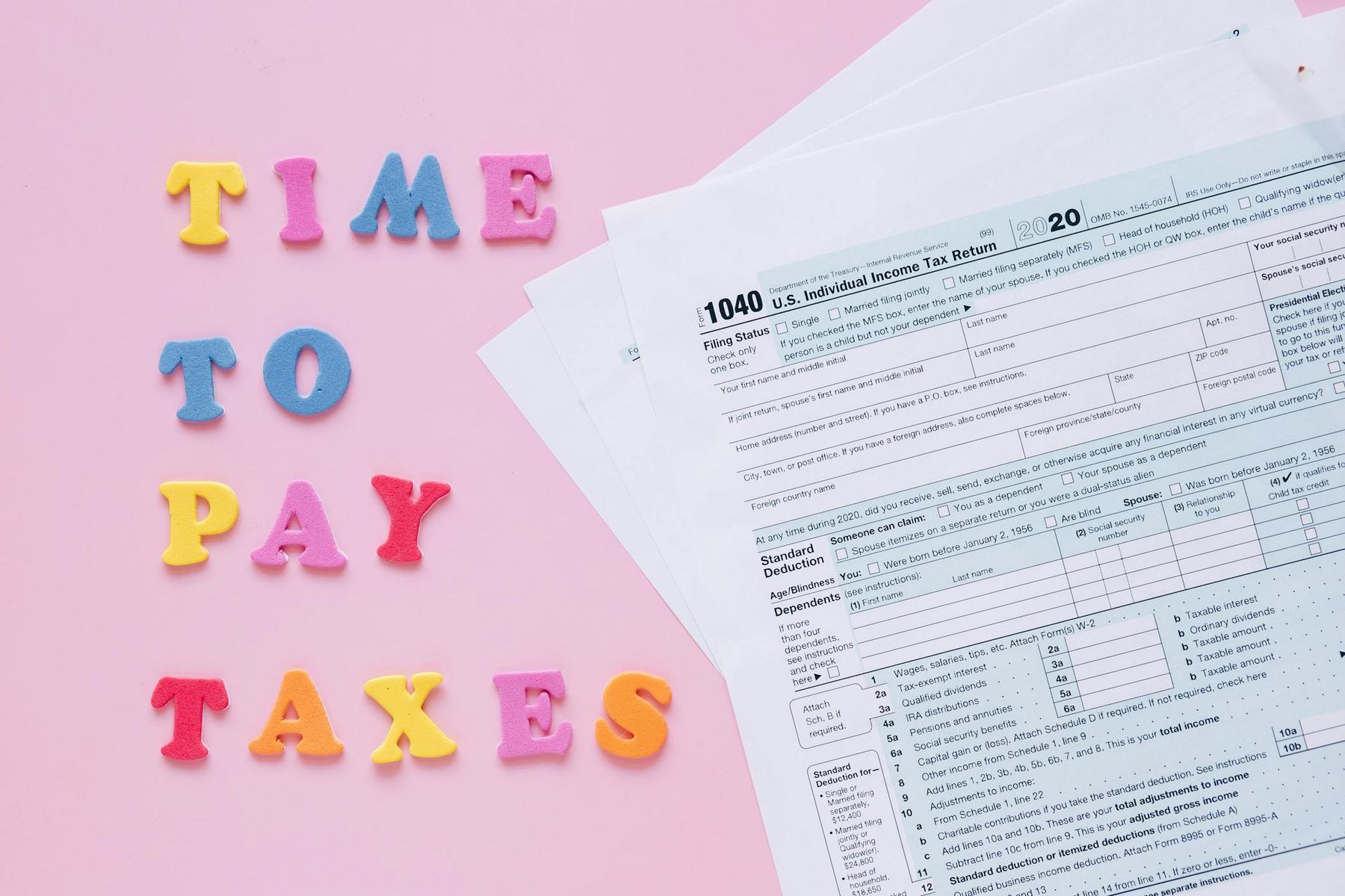Colorful letters spelling "Time to Pay Taxes" beside U.S. 1040 tax forms on a pink background.