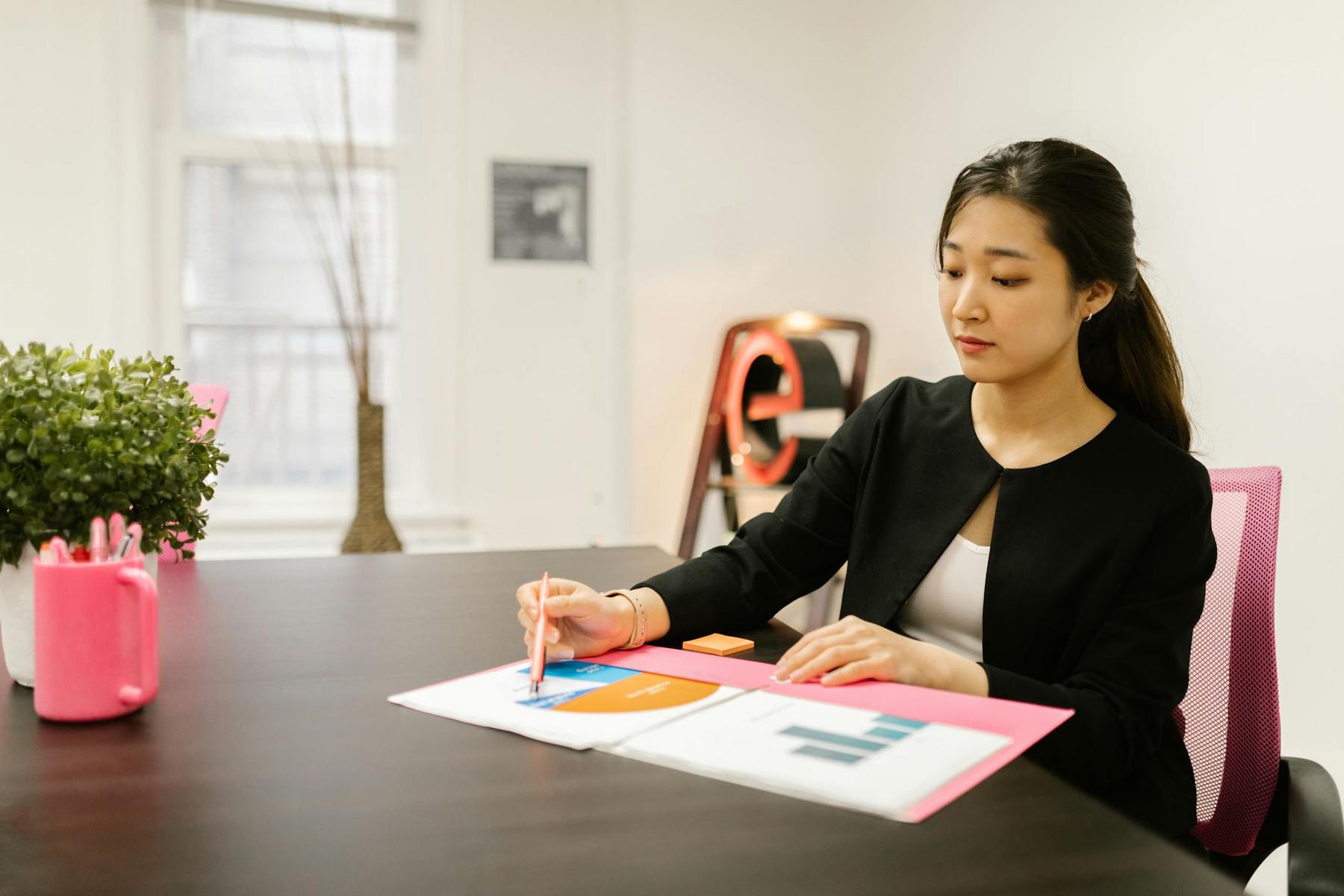 A young Asian woman in a black blazer sits at a desk, looking at charts and graphs.