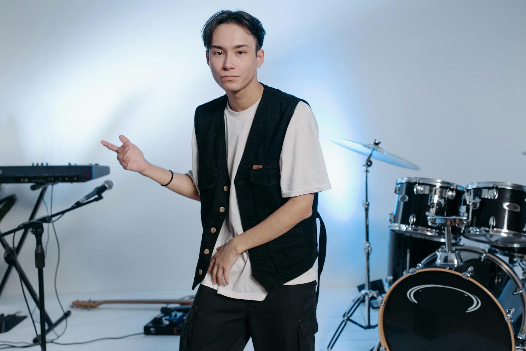 A young man in a black vest and white t-shirt stands in a music studio, gesturing with one hand. Musical instruments, including drums and a keyboard, are visible in the background.