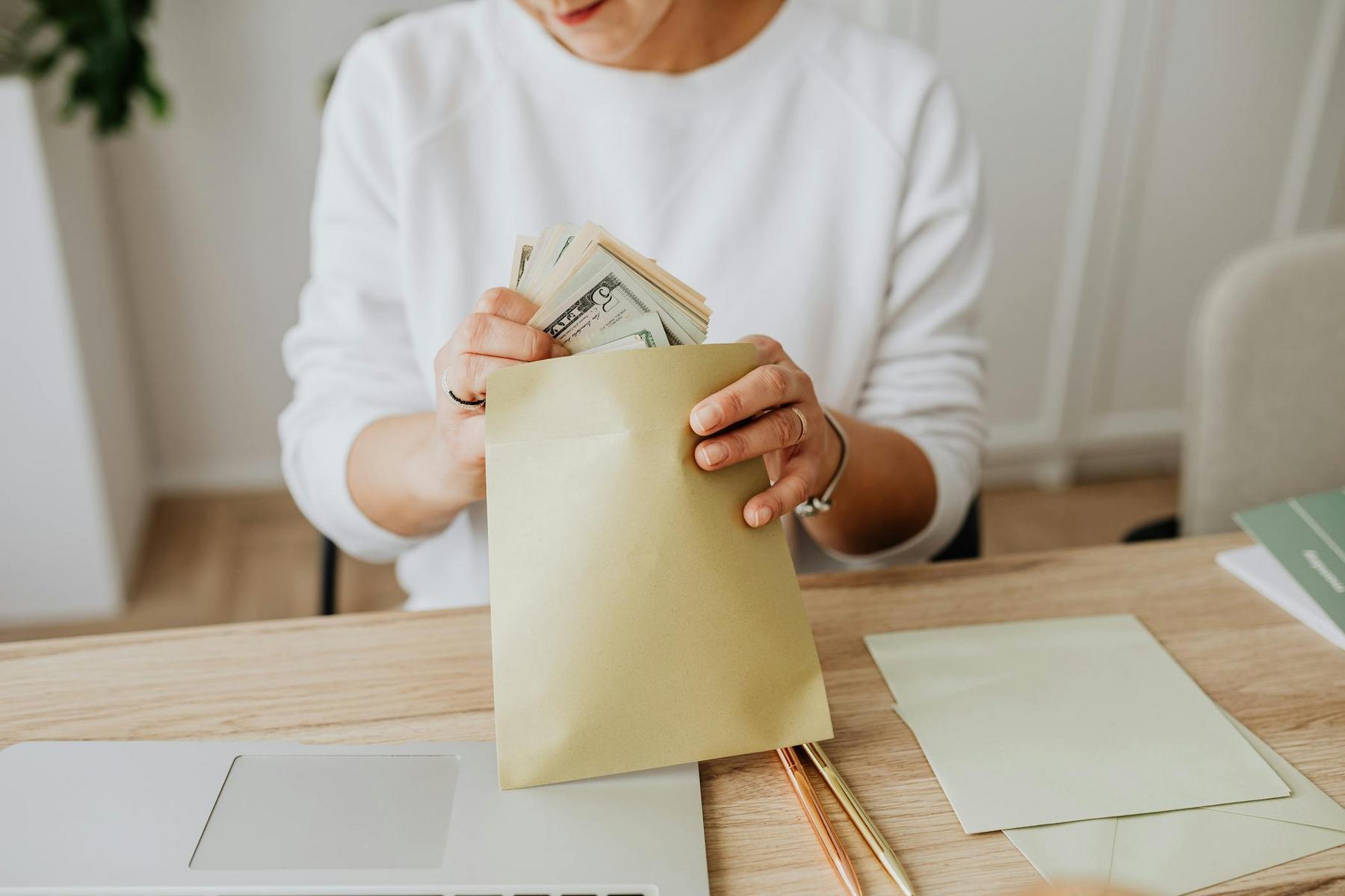 Person placing a stack of US dollar bills into a brown envelope while sitting at a desk with a laptop and papers.