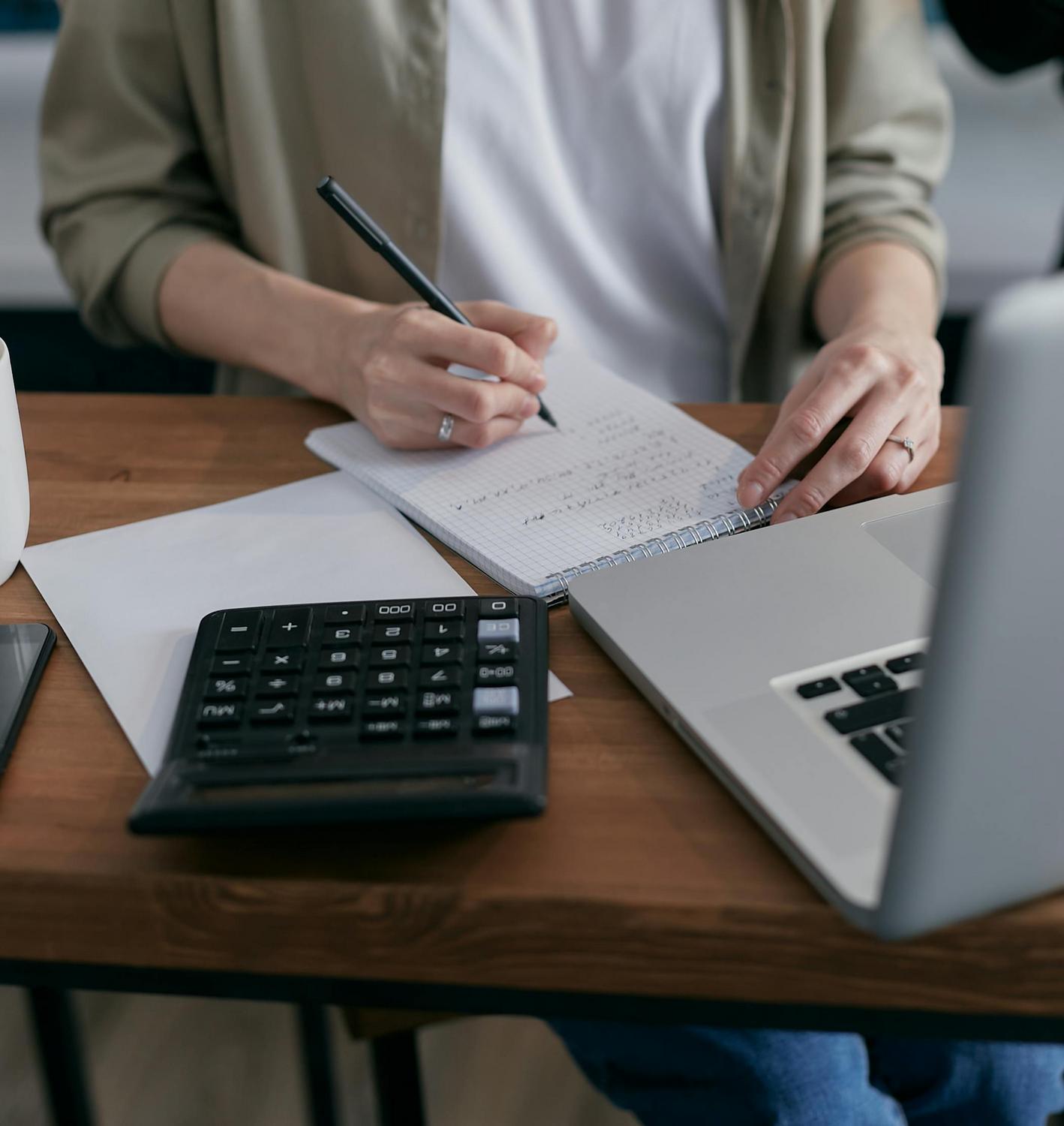 Person writing in a notebook next to a laptop and calculator on a wooden desk, with paperwork and office supplies visible.
