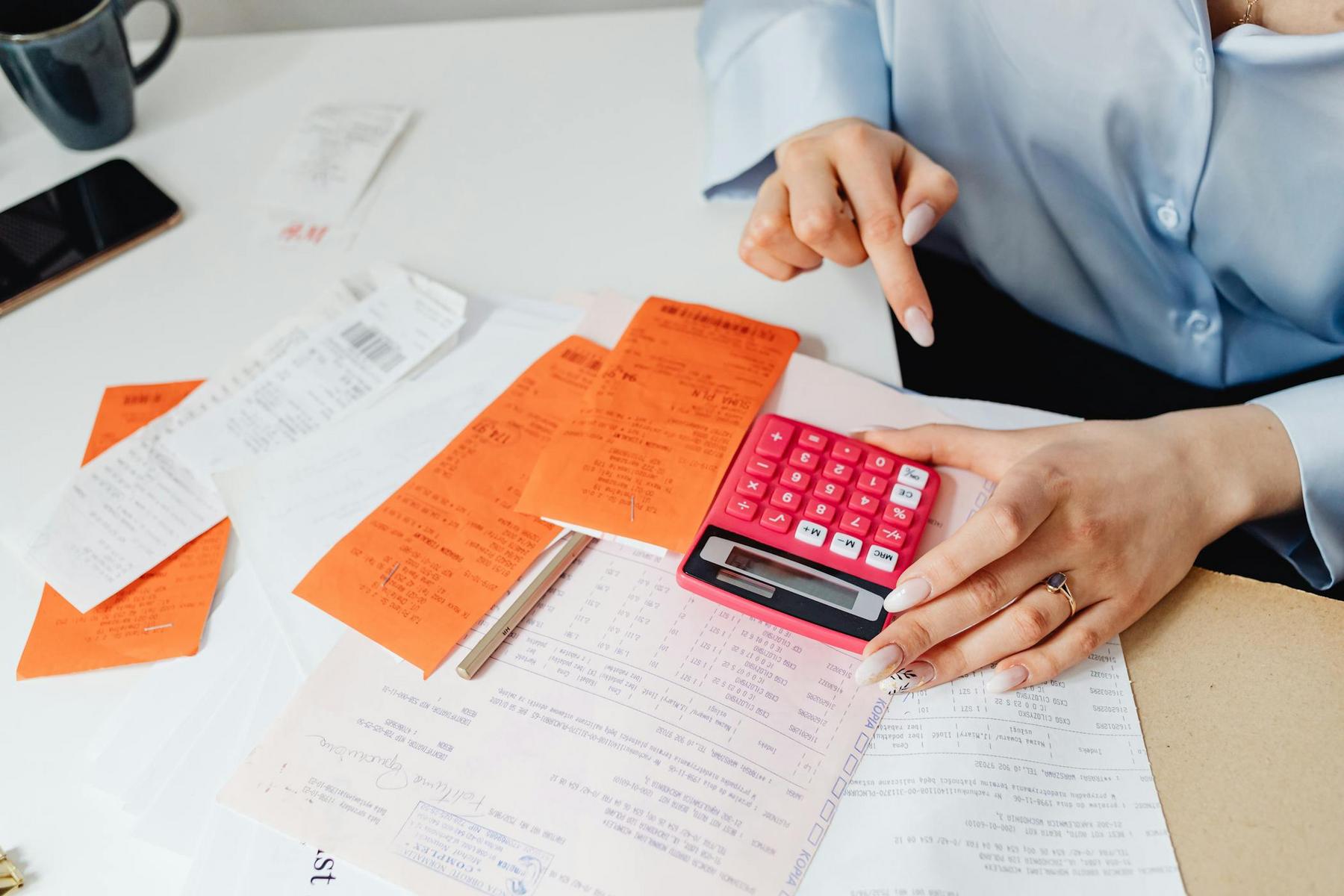 Person using a red calculator and reviewing bills, receipts, and documents at a desk with a pen and coffee mug nearby.