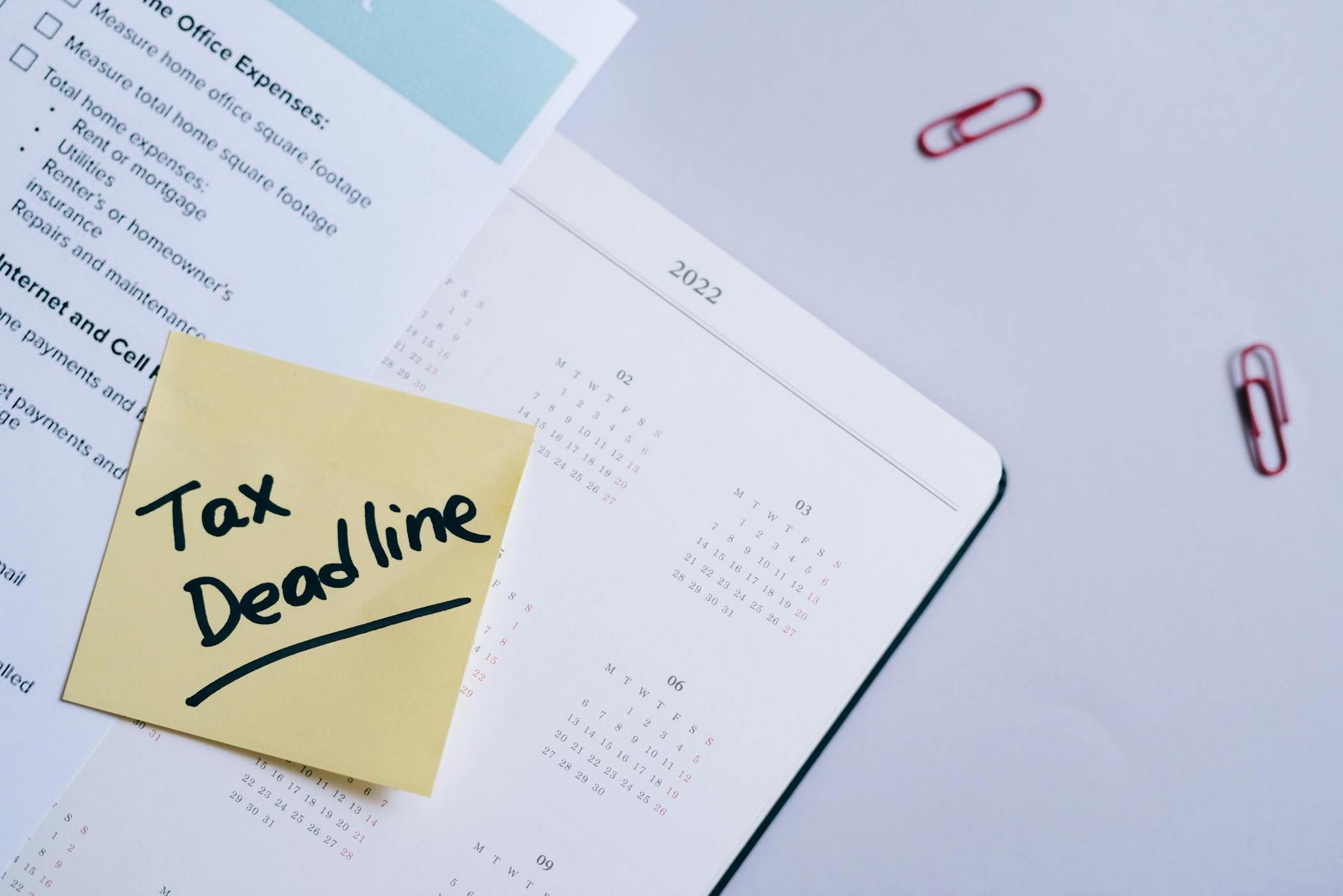 A sticky note with "Tax Deadline" written on it sits on top of a calendar, alongside documents and red paper clips.
