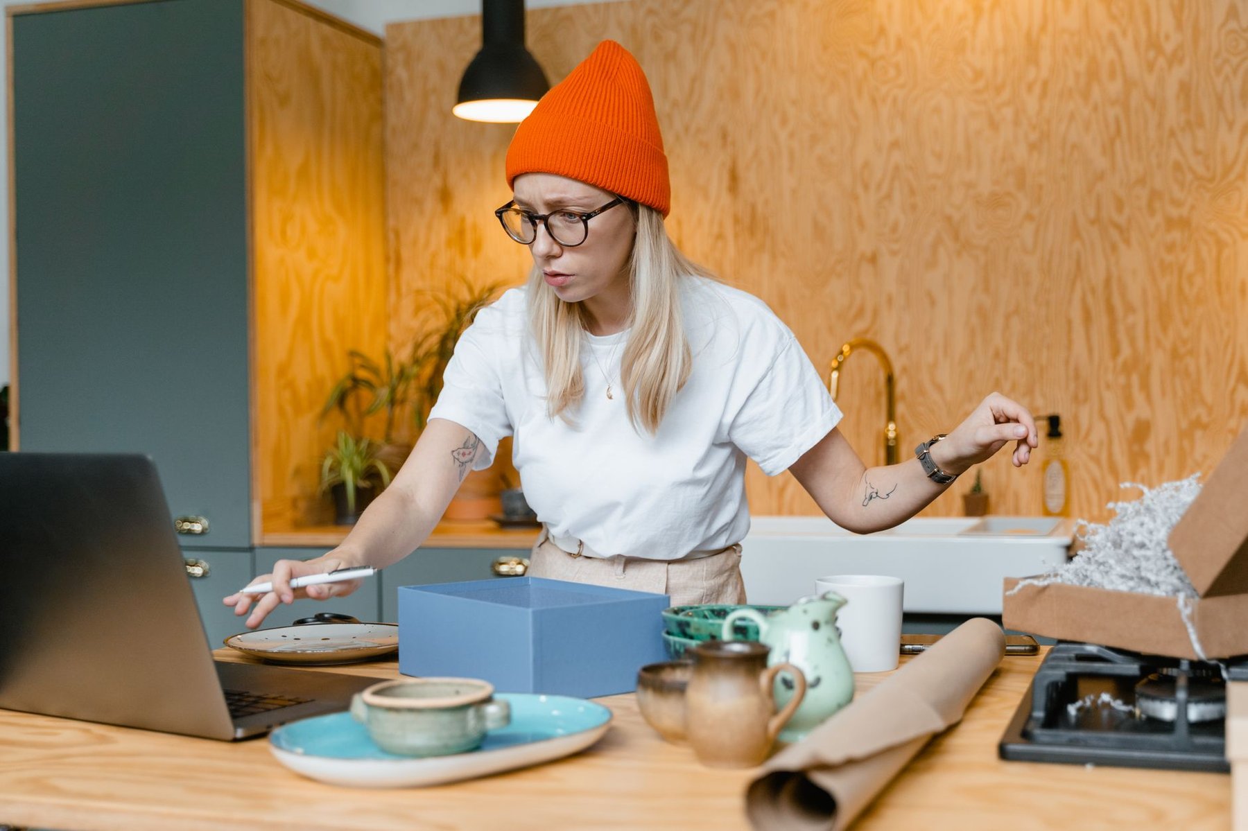 Ceramic artist packing handmade pottery orders at her kitchen workbench while checking her online shop on a laptop