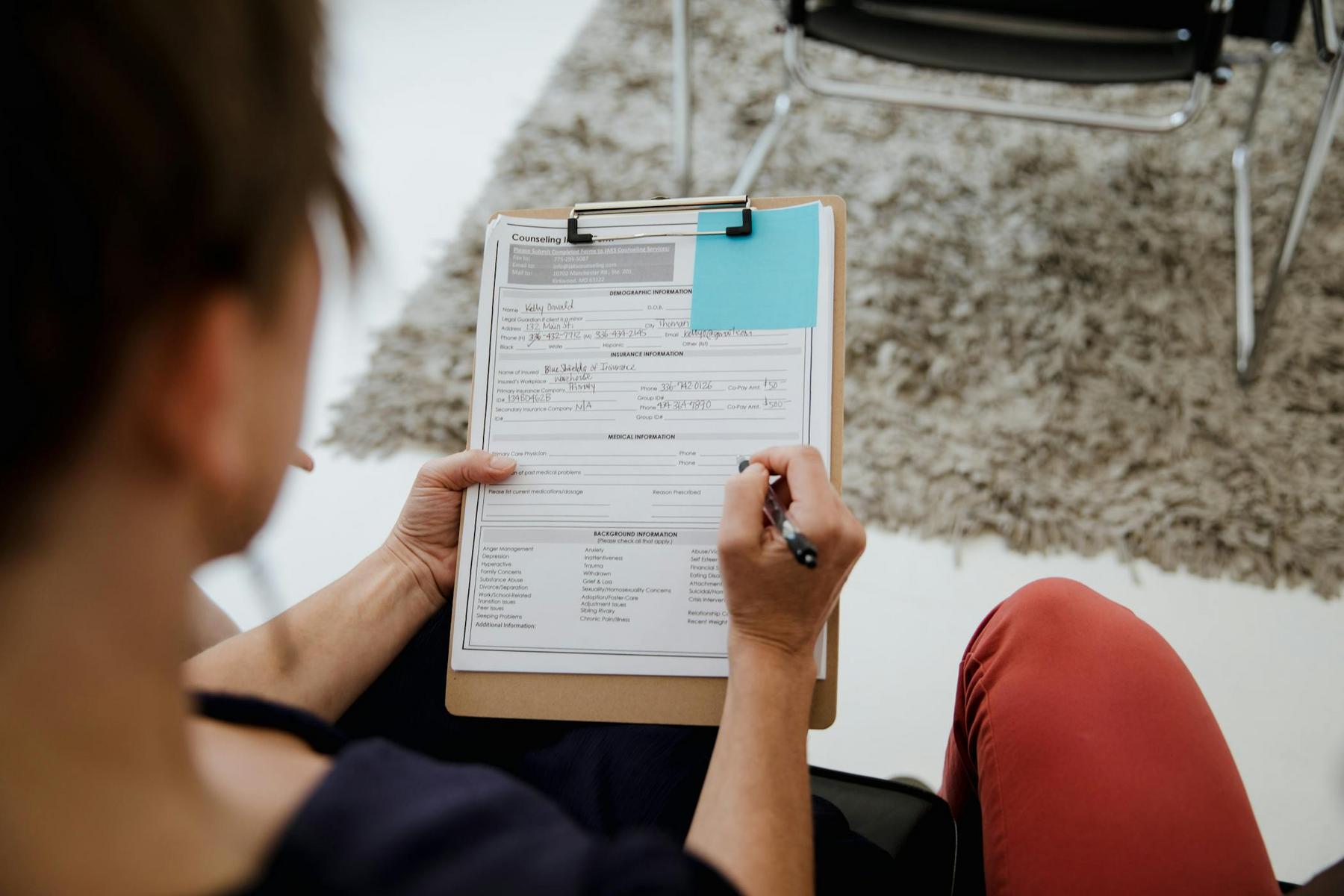 A person holding a clipboard fills out a counseling intake form with a pen; a chair and a rug are visible in the background.