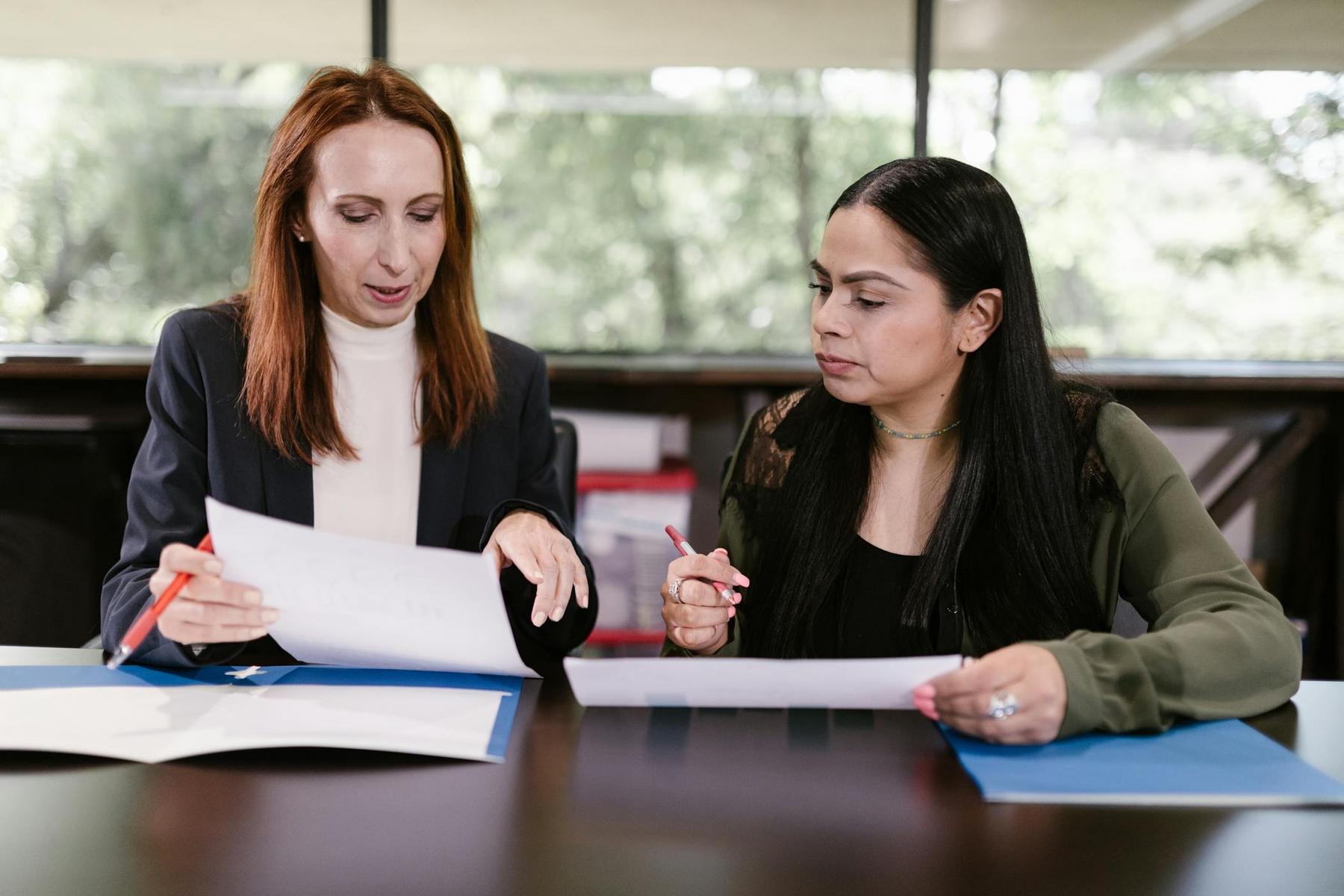 Two women sit at a table, reviewing and discussing documents together in a professional office setting.