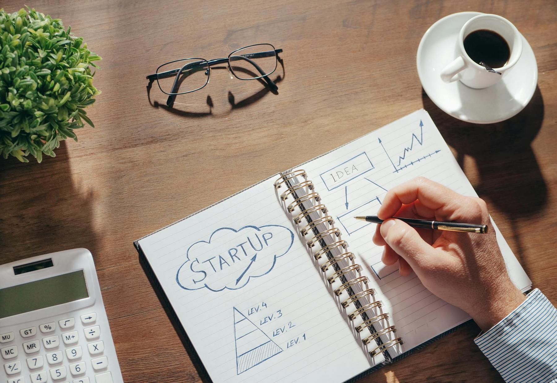 A hand drawing startup-related charts and notes in a notebook on a desk with a calculator, glasses, a potted plant, and a cup of coffee.