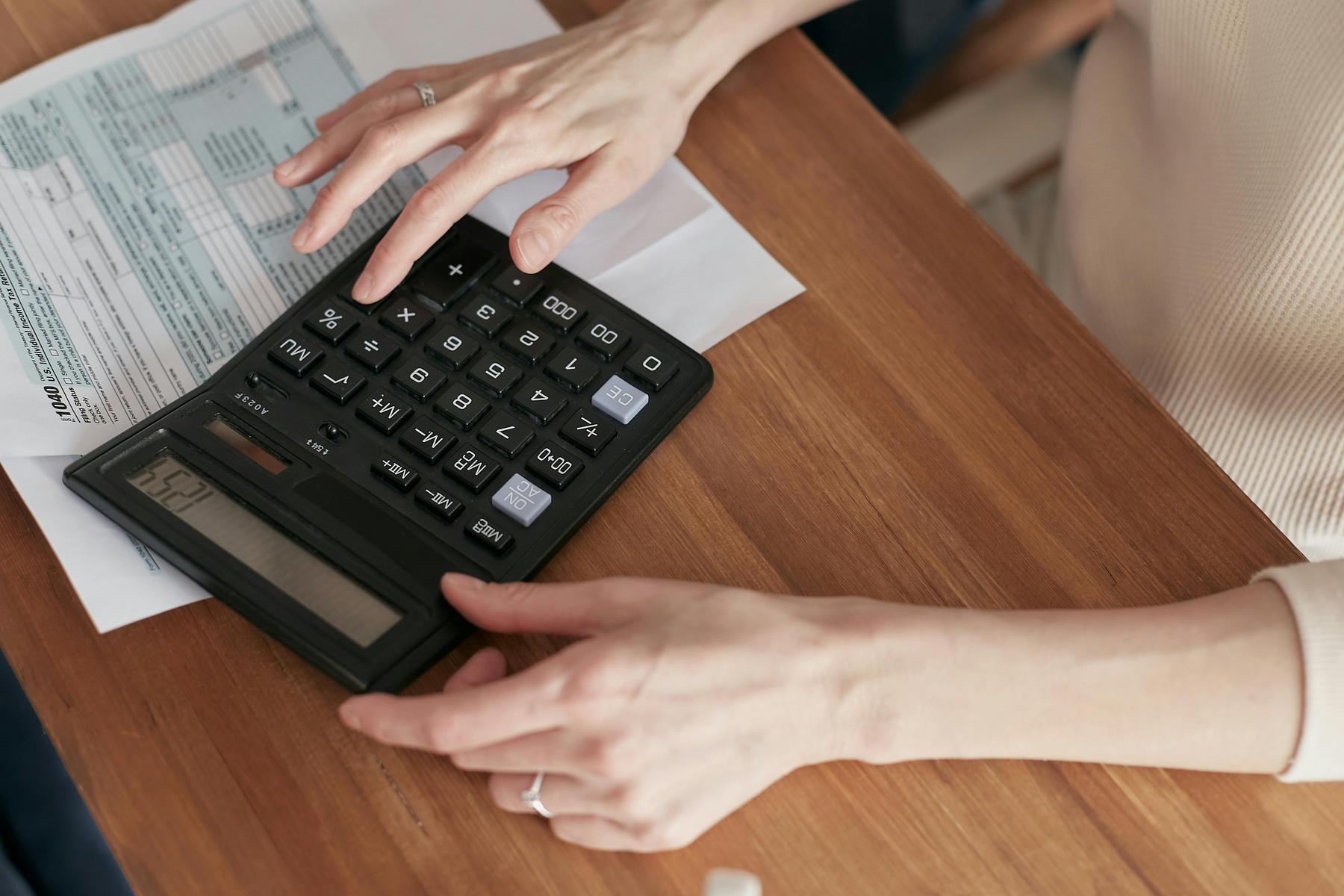 A person uses a large calculator on a wooden table with papers, possibly tax or financial documents, visible underneath.