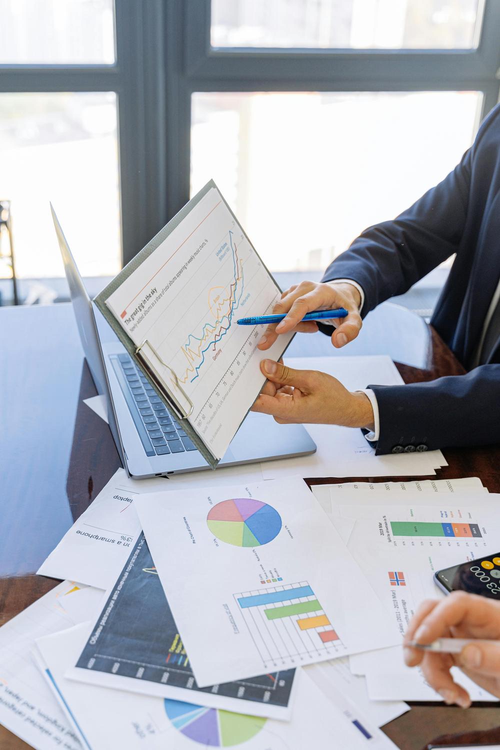 Two people review financial charts and graphs on printed sheets and a clipboard, analyzing data with pens in a brightly lit office setting.