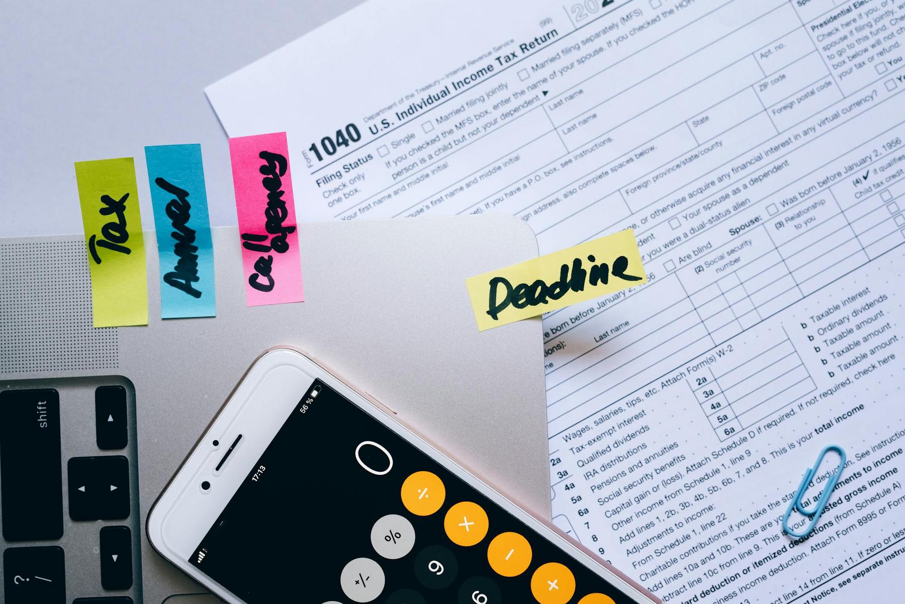 1040 tax form, sticky notes labeled "Tax," "Answer," "Call Jenny," and "Deadline," a smartphone calculator, part of a laptop, and paper clips on a desk.