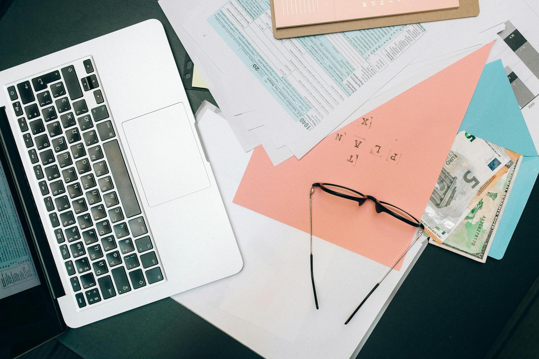 A laptop next to scattered documents, eyeglasses, and a pink folder on a desk.
