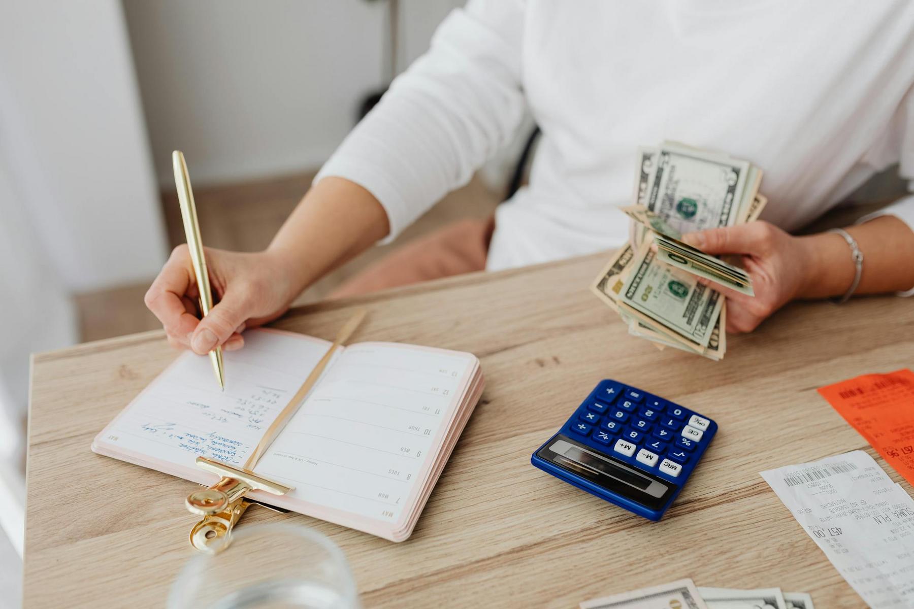 Person writing in a notebook while holding cash, with a calculator and receipts on the wooden desk.