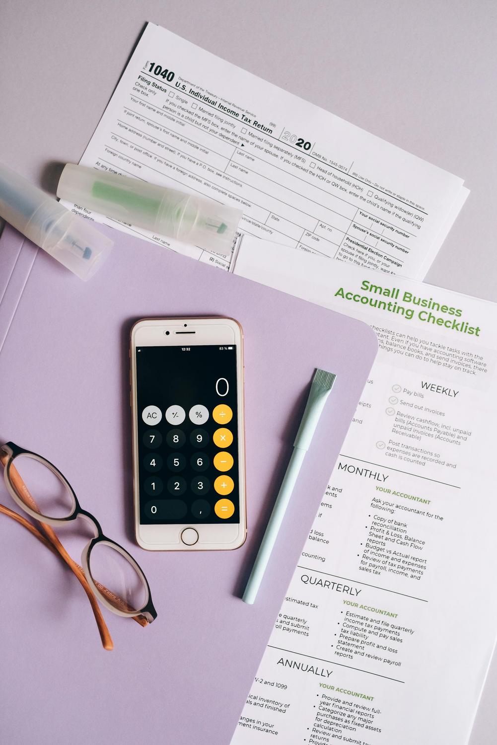 A smartphone with a calculator app, glasses, a pen, and documents including a 1040 tax form and a small business accounting checklist on a desk.