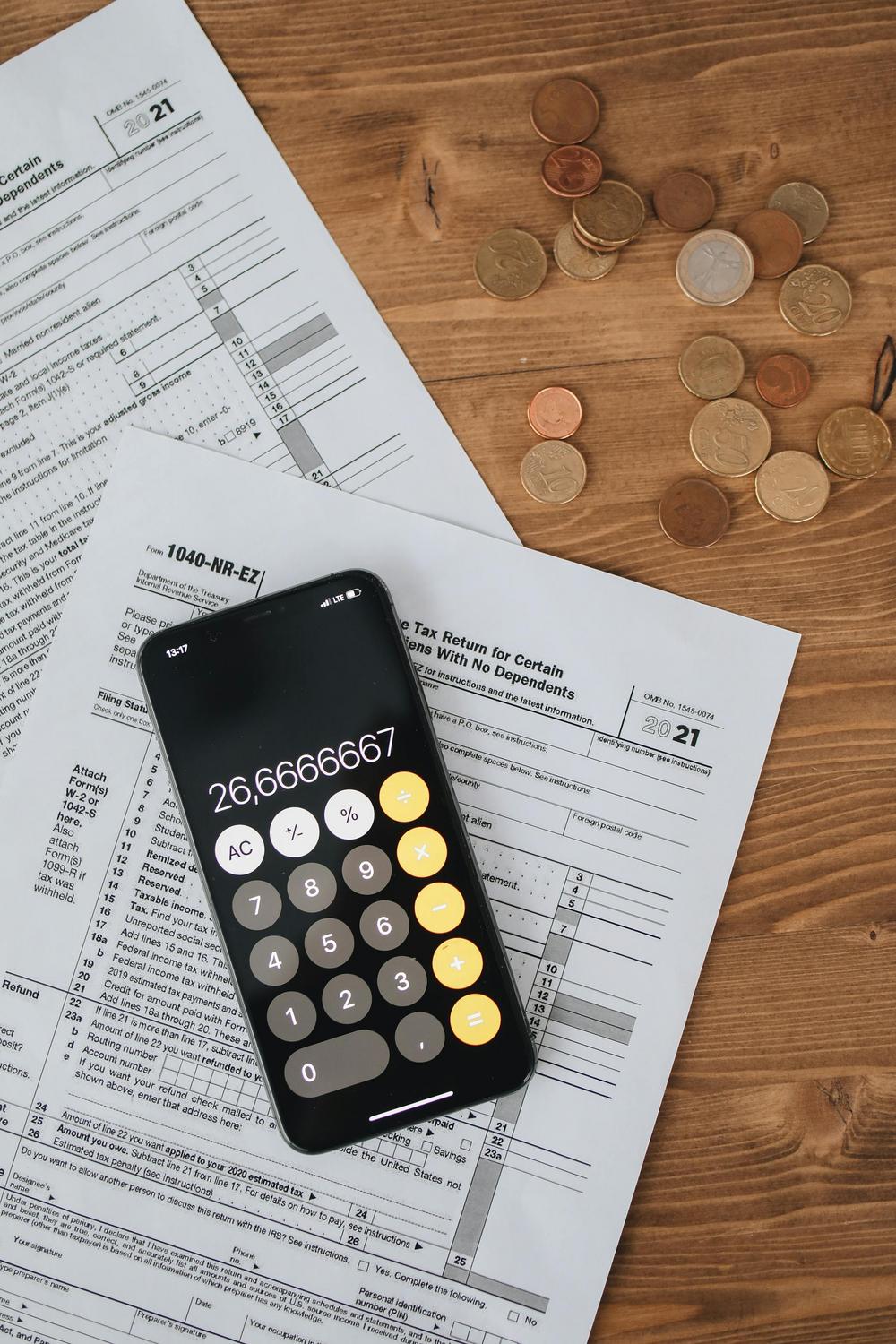 A smartphone with a calculator app, tax forms, and scattered coins are placed on a wooden surface.