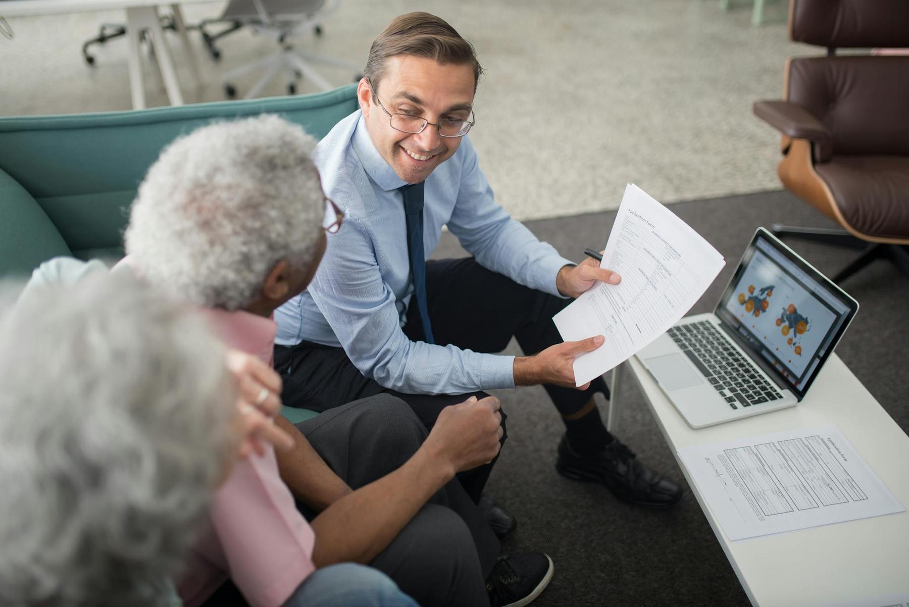A man in business attire shows paperwork to two older adults on a sofa, with a laptop displaying graphs on a coffee table.