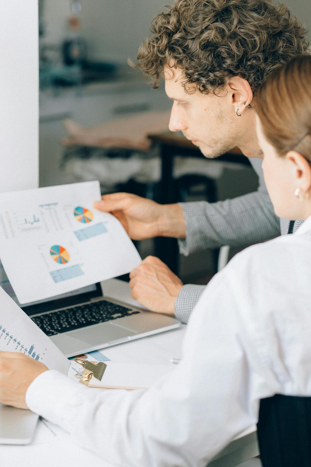 Two people review printed charts and graphs at a desk, with a laptop open in front of them and a clipboard nearby.