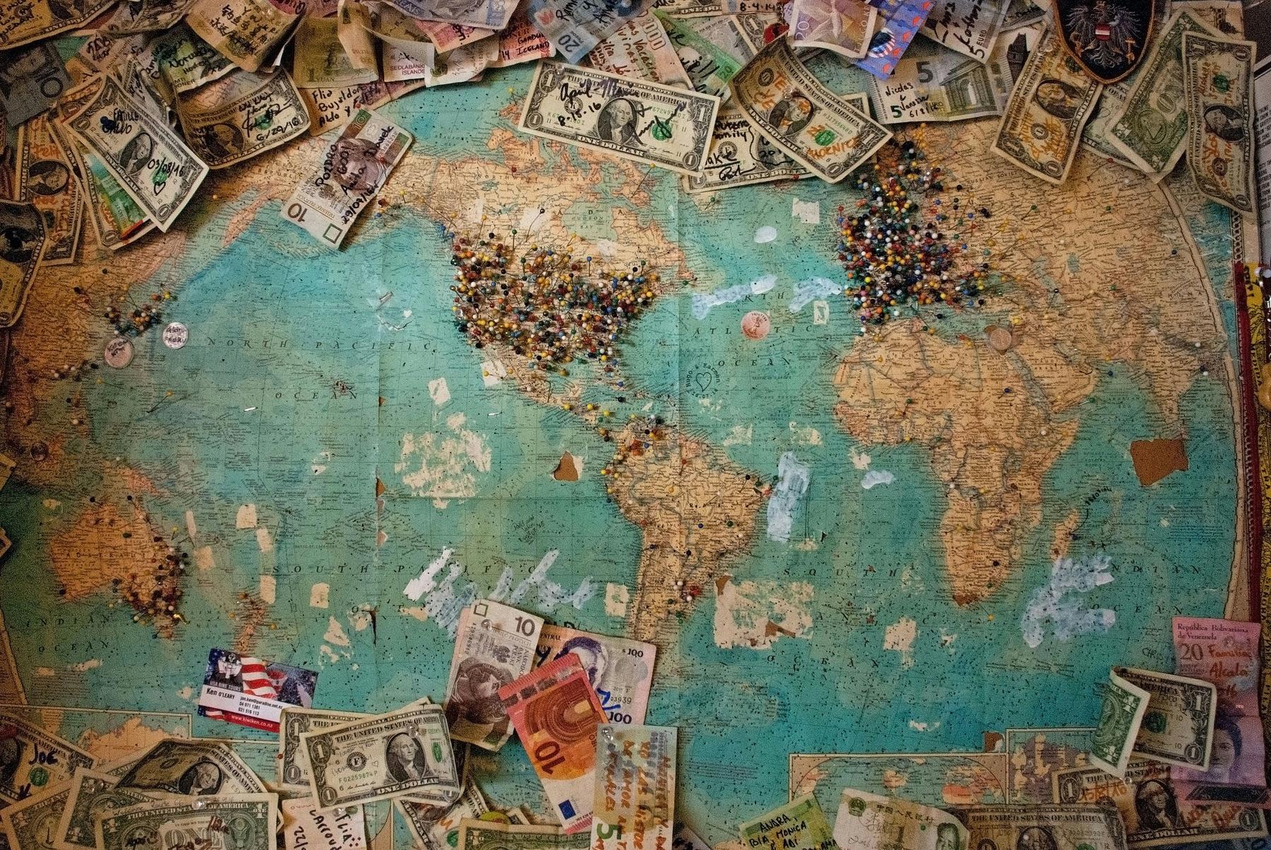 A world map with many pins marking locations, surrounded by various international currency notes.