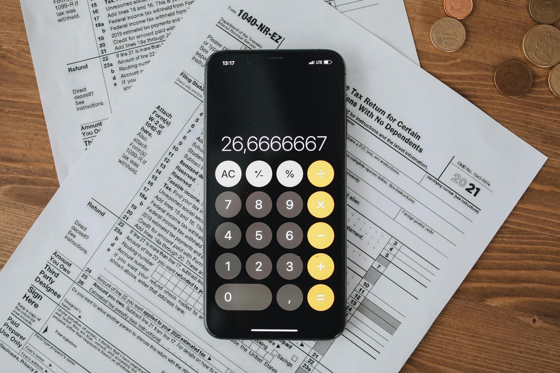 A smartphone displaying a calculator app with the number 26,6666667 rests on tax documents, with coins scattered on a wooden surface nearby.