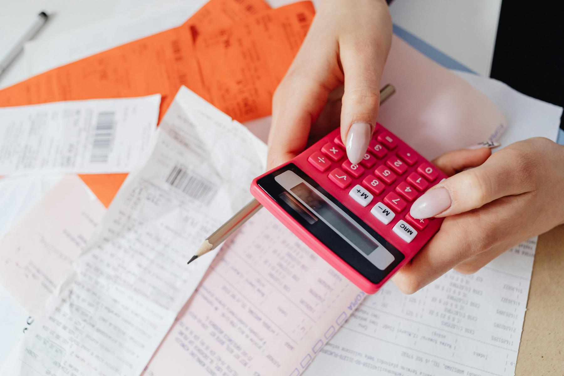 Person using a pink calculator with receipts, bills, and documents spread on a table, holding a pencil in one hand.