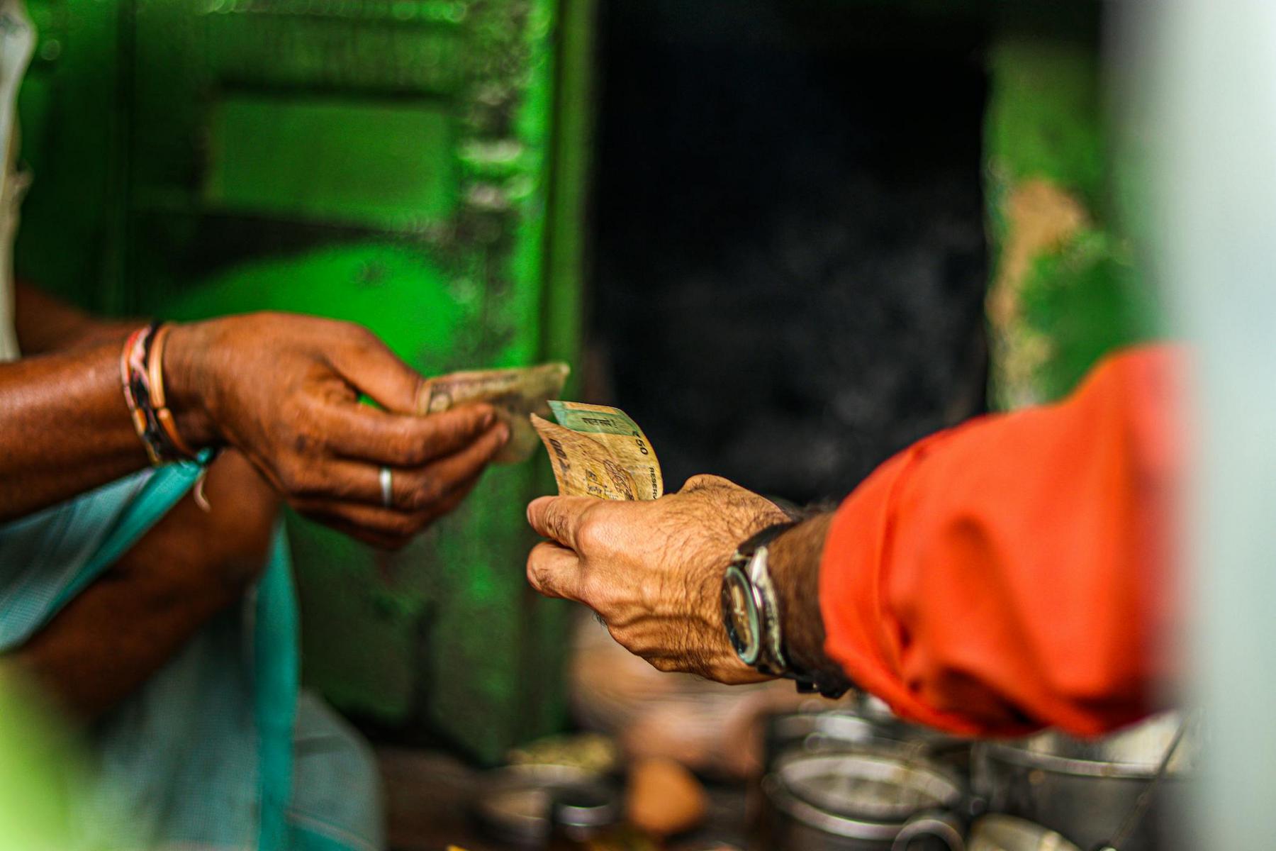 Two people exchange Indian currency notes, with one hand giving and the other receiving money, against a green background.
