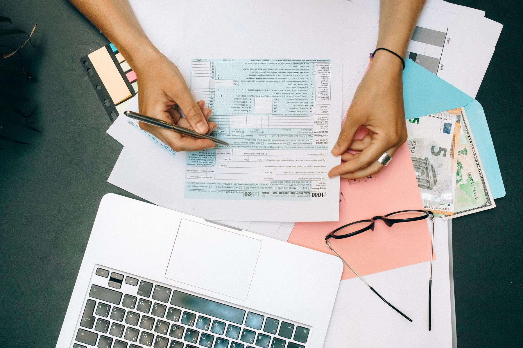 A person fills out a tax form at a desk with a laptop, glasses, documents, cash, and sticky notes.