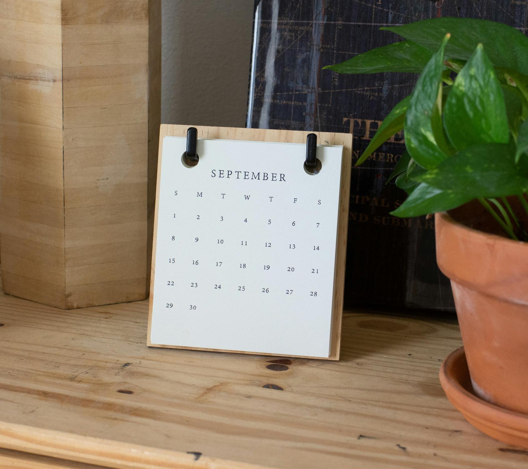 A wooden desk calendar displaying the month of September is placed on a wooden shelf next to a potted plant and a book.