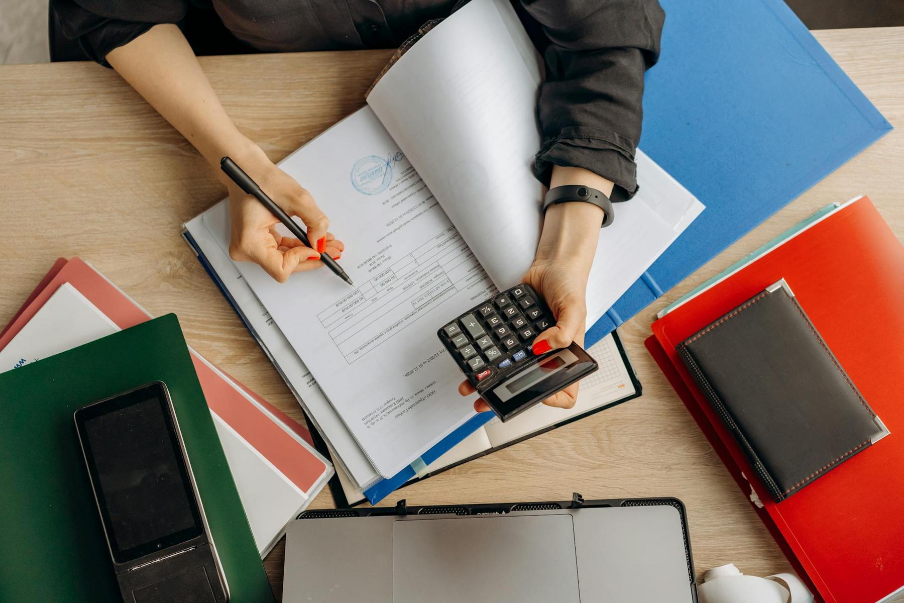 Person using a calculator and writing on a document at a desk with folders, notebooks, and a smartphone.