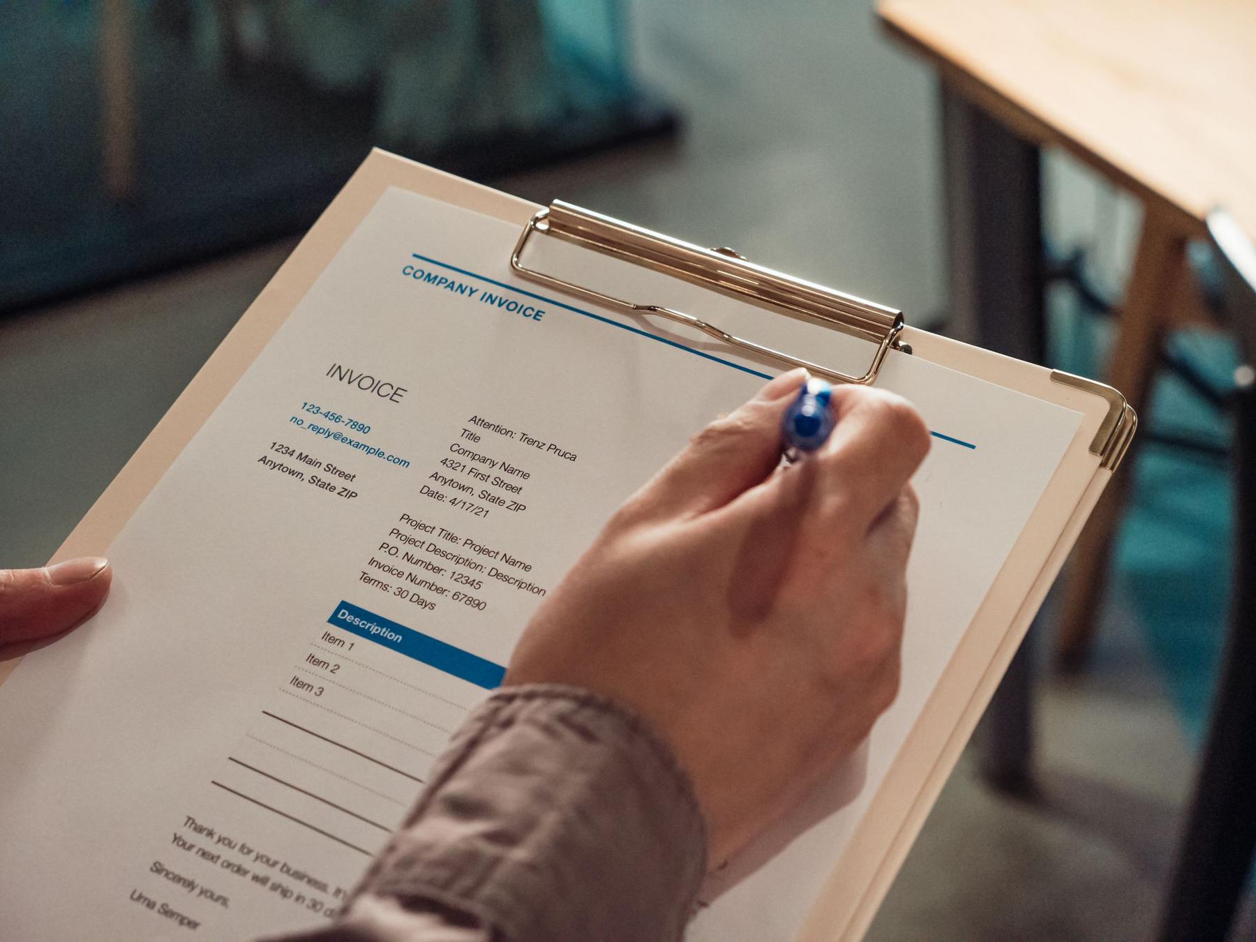 A person holds a clipboard with an invoice document, using a pen to point at or review the information on the page.