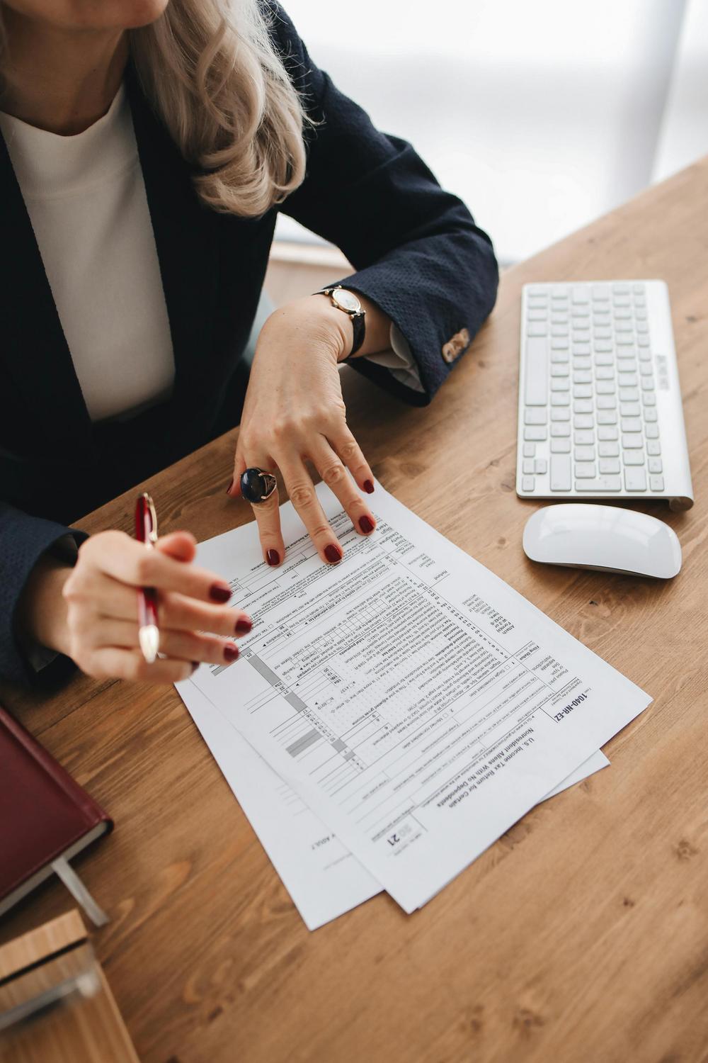 A person in business attire reviews and marks documents at a wooden desk with a computer keyboard and mouse.