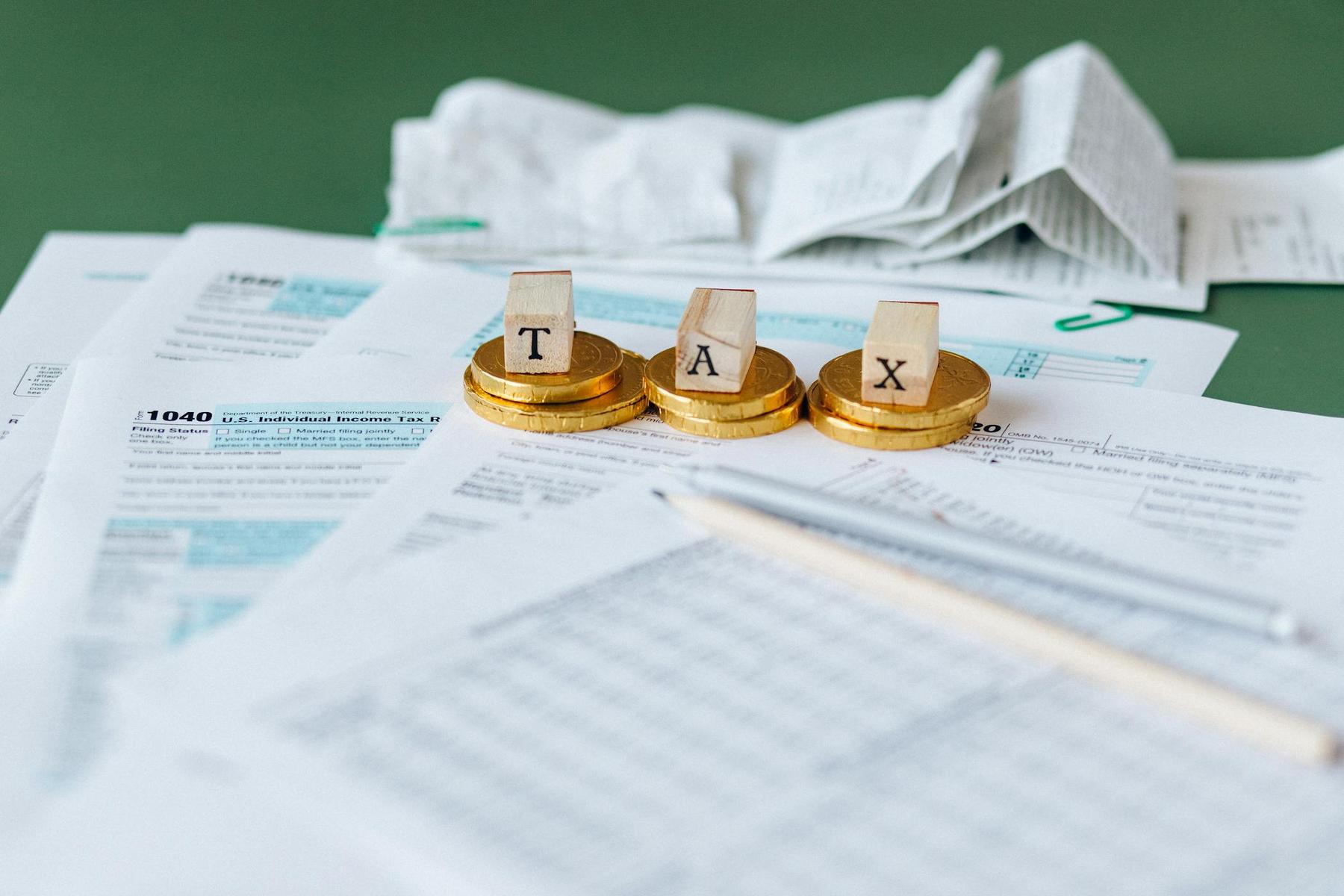 Tax forms, receipts, a pencil, and gold coins with letter blocks spelling "TAX" are arranged on a table.