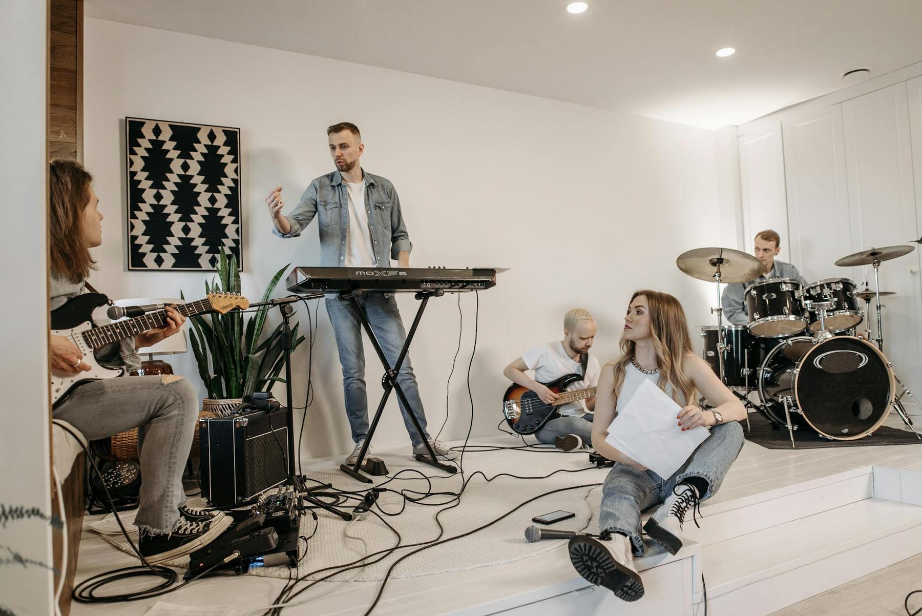 A five-member band rehearses indoors; one person stands at a keyboard, two play guitar, one plays drums, and one sits on the floor holding sheet music.