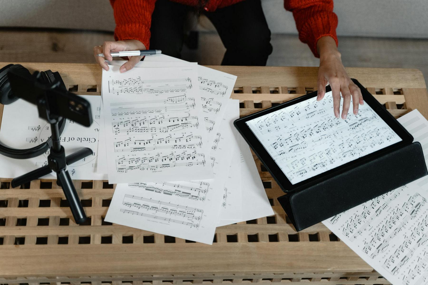 Person arranges printed sheet music and a tablet displaying sheet music on a wooden table, with a tripod and headphones also present.