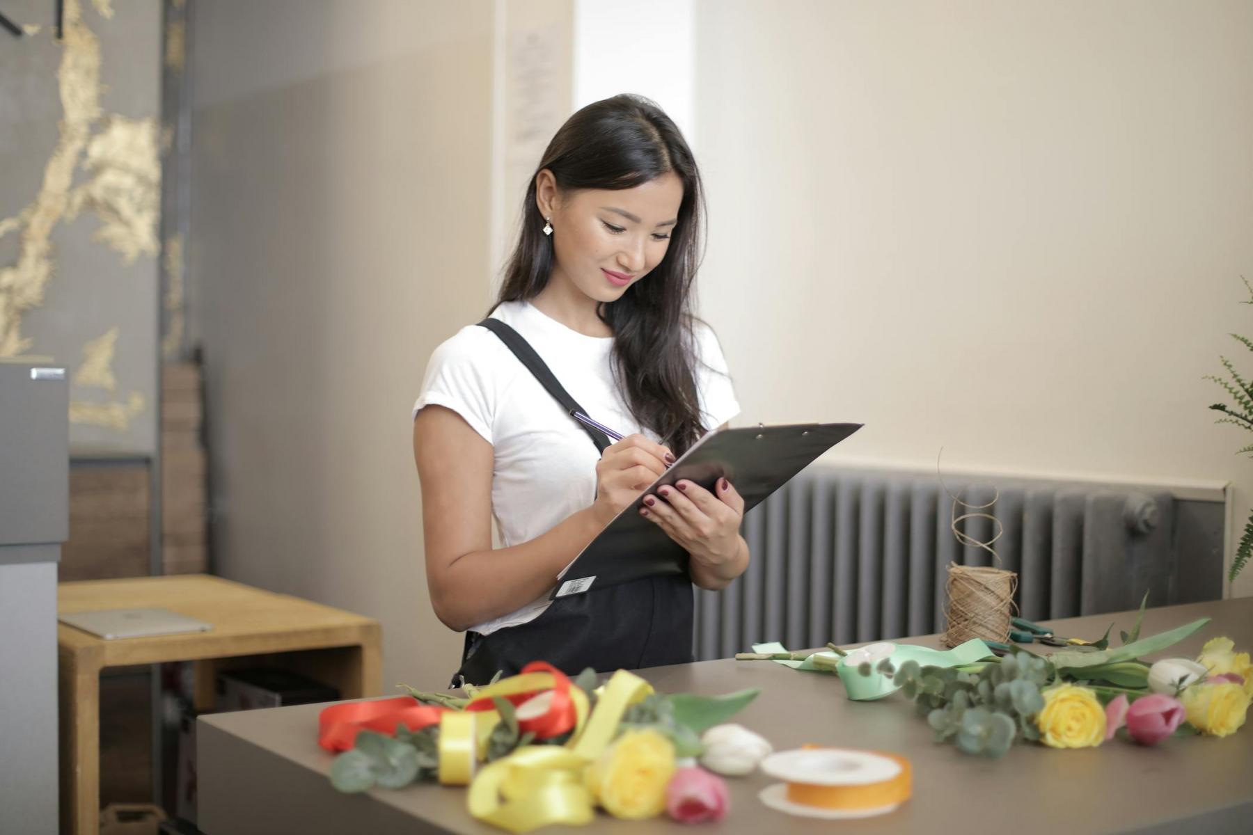 A woman in a white shirt and black apron writes on a clipboard at a counter with flowers, ribbons, and craft supplies.