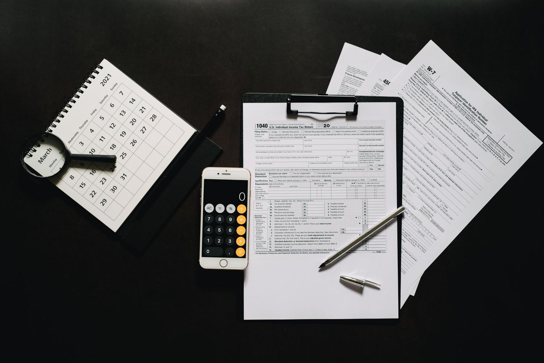 A clipboard with tax forms, a pen, a smartphone with a calculator app open, papers, a calendar, a magnifying glass, and a pencil arranged on a dark desk.