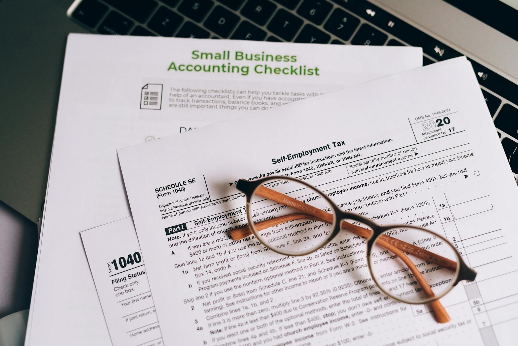 A pair of glasses rests on top of tax forms and a small business accounting checklist, with part of a laptop keyboard visible in the background.