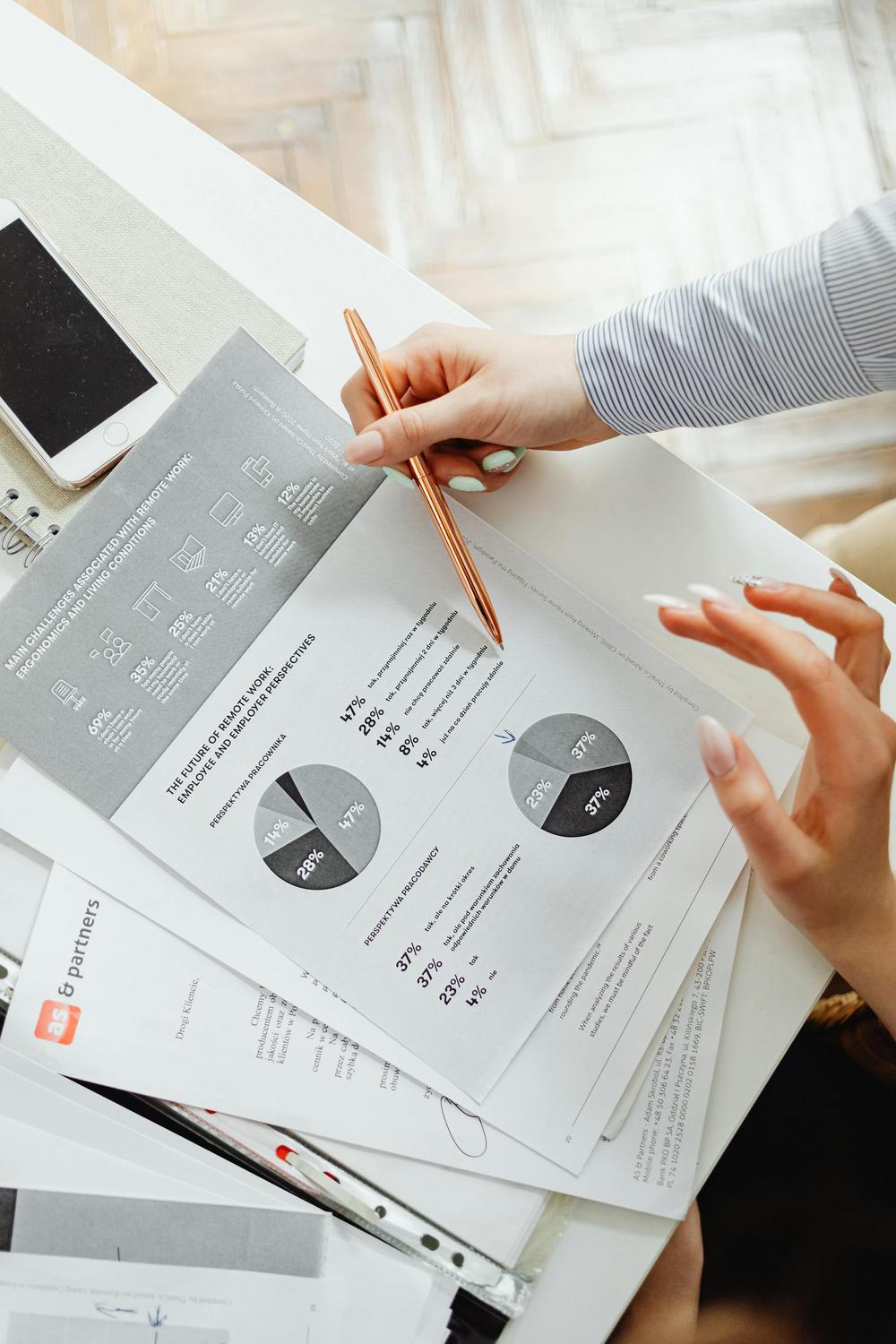 Person holding a pen and pointing at a printed report with pie charts and statistics on a desk with documents and a smartphone.
