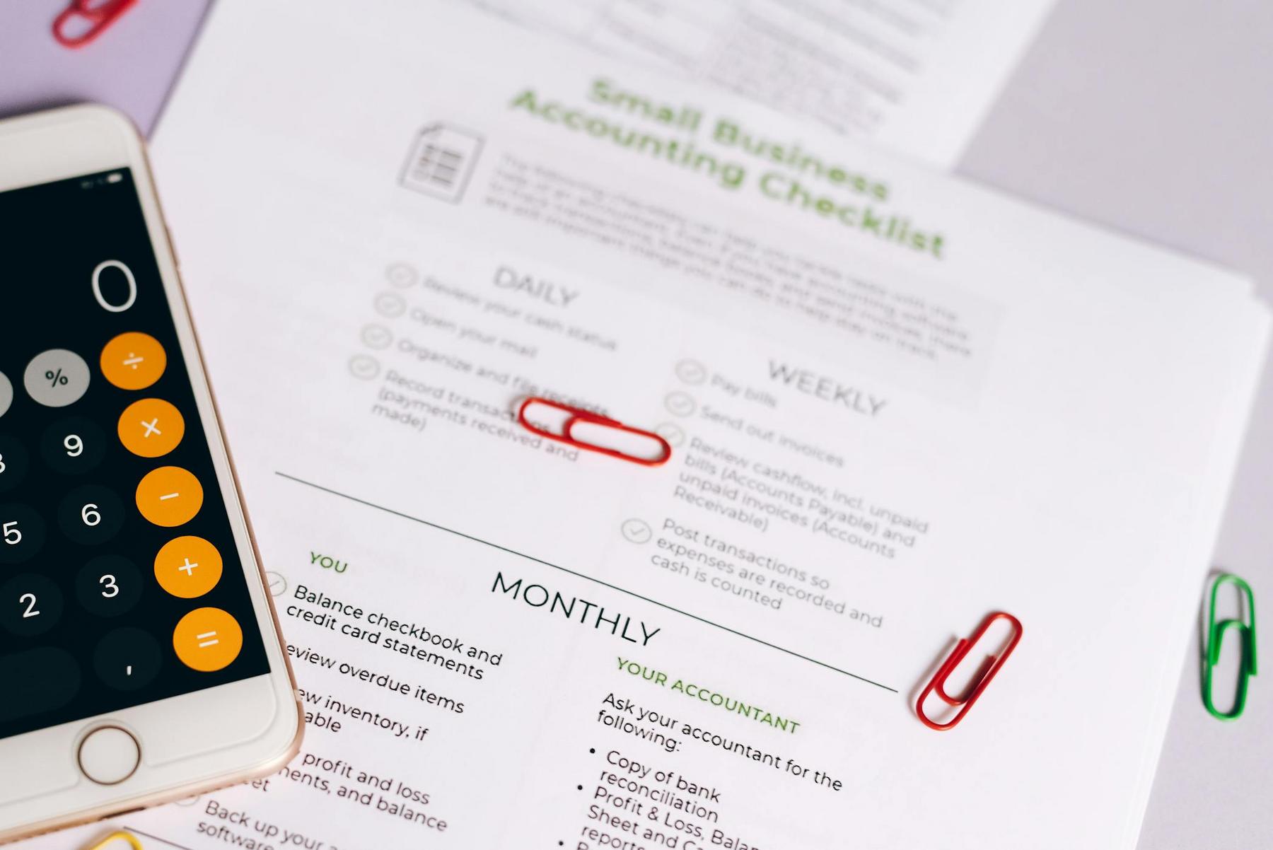 A smartphone with a calculator app, accounting documents titled "Small Business Accounting Checklist," and red paperclips on a desk.