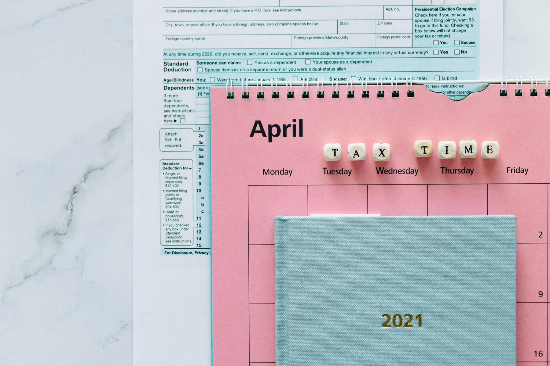 A tax form, a desk calendar open to April, and a blue 2021 planner. Letter blocks spell "TAX TIME" across the calendar.