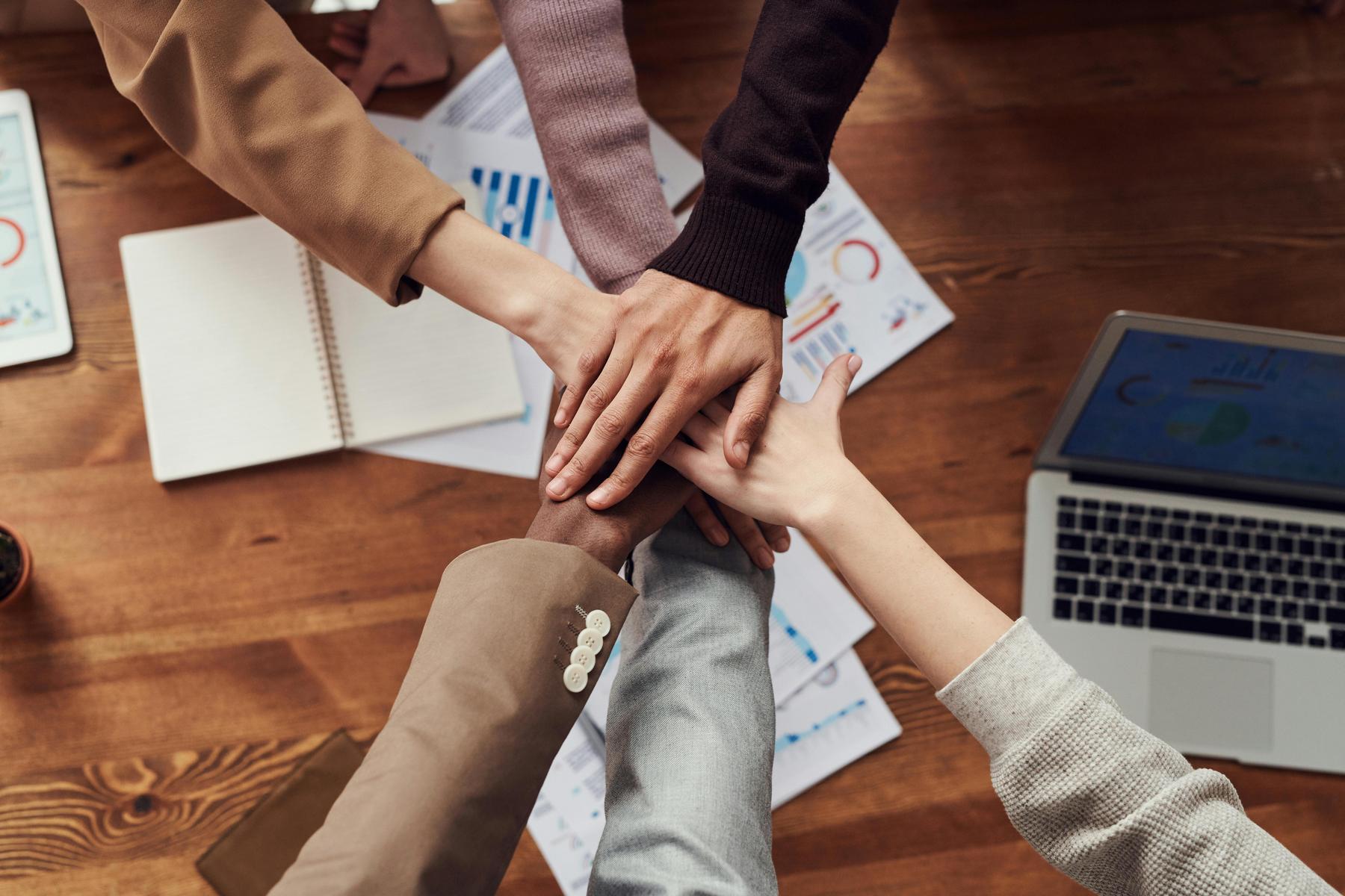 Five people place their hands together in a stack over a wooden table with documents, graphs, a notebook, and a laptop.