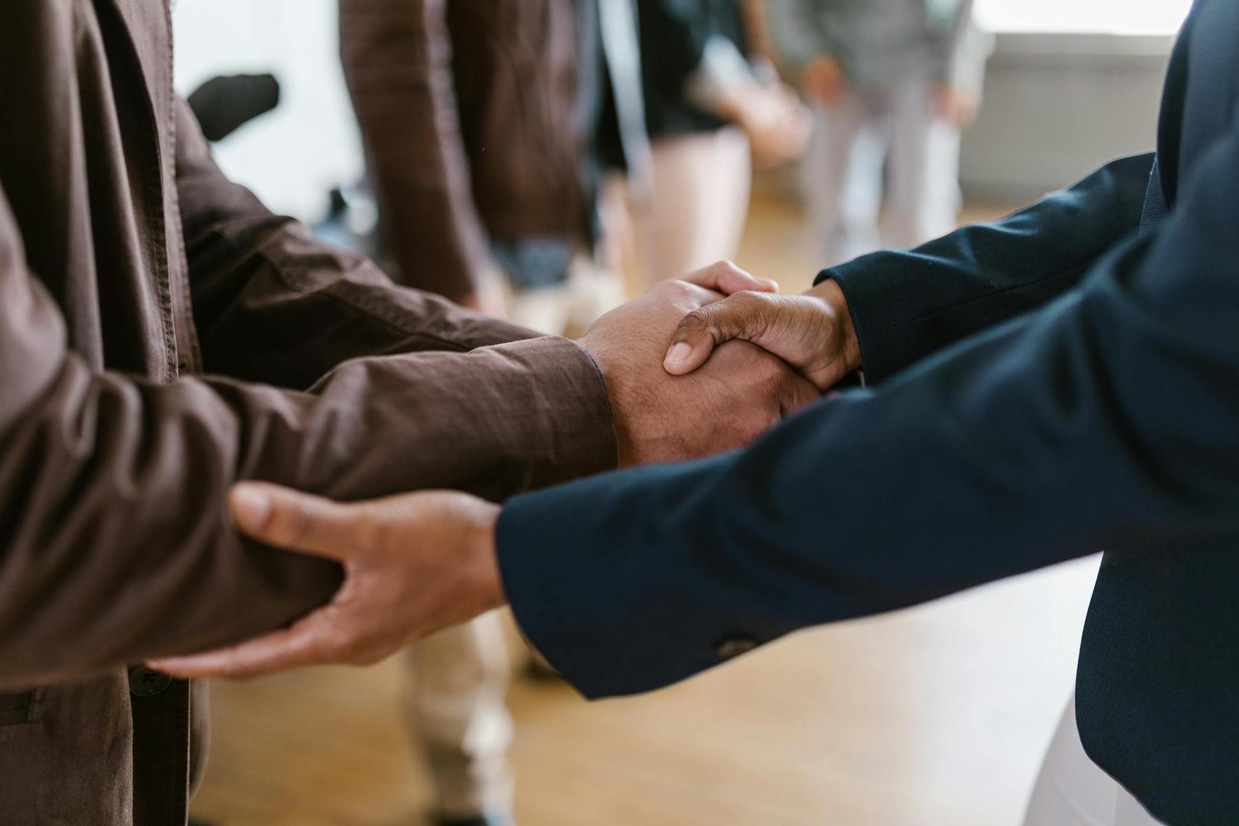 Two people shaking hands in a business setting, with others blurred in the background.