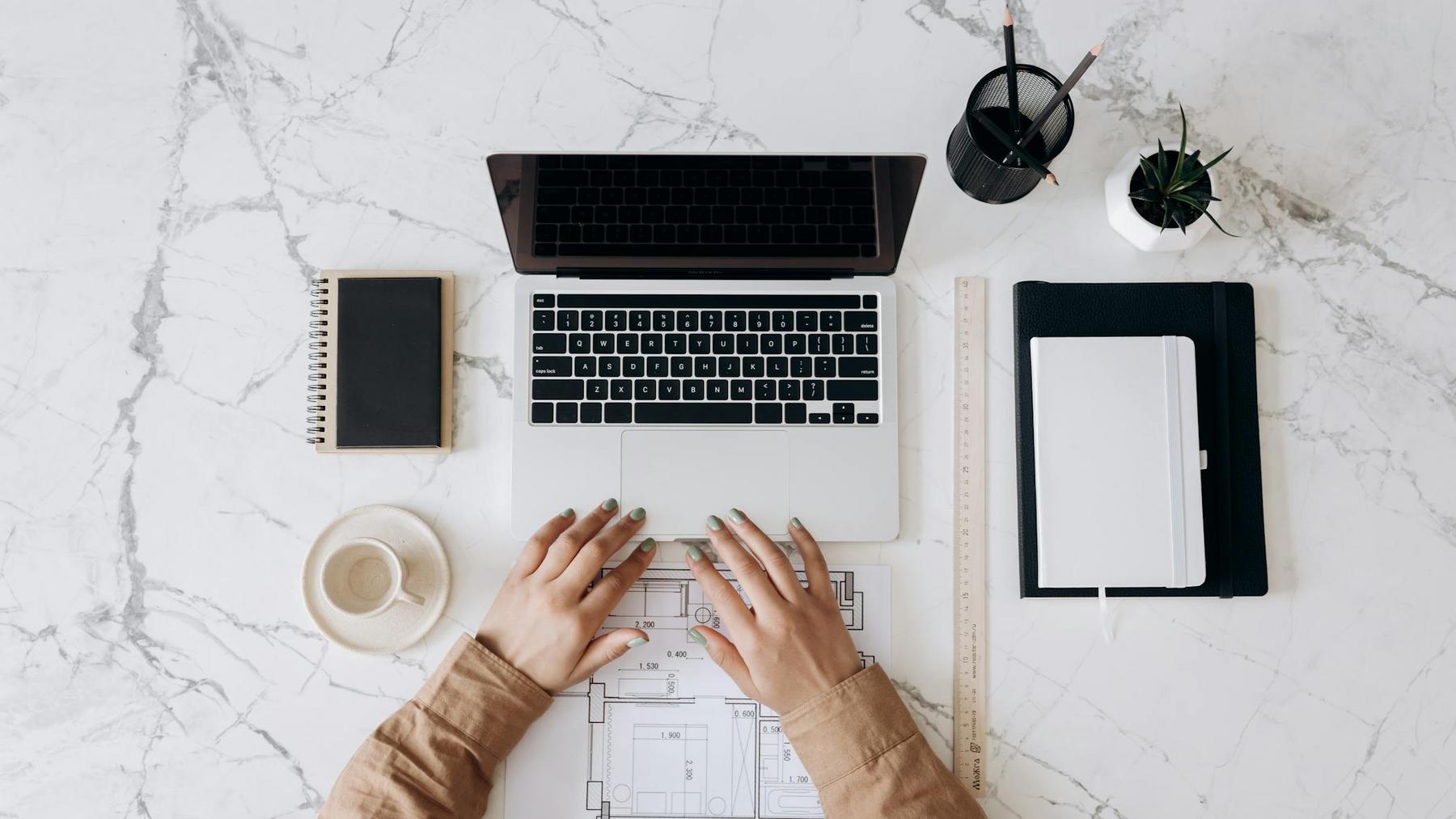 A person types on a laptop at a marble desk with a notebook, coffee cup, ruler, pen holders, a potted plant, and architectural plans.