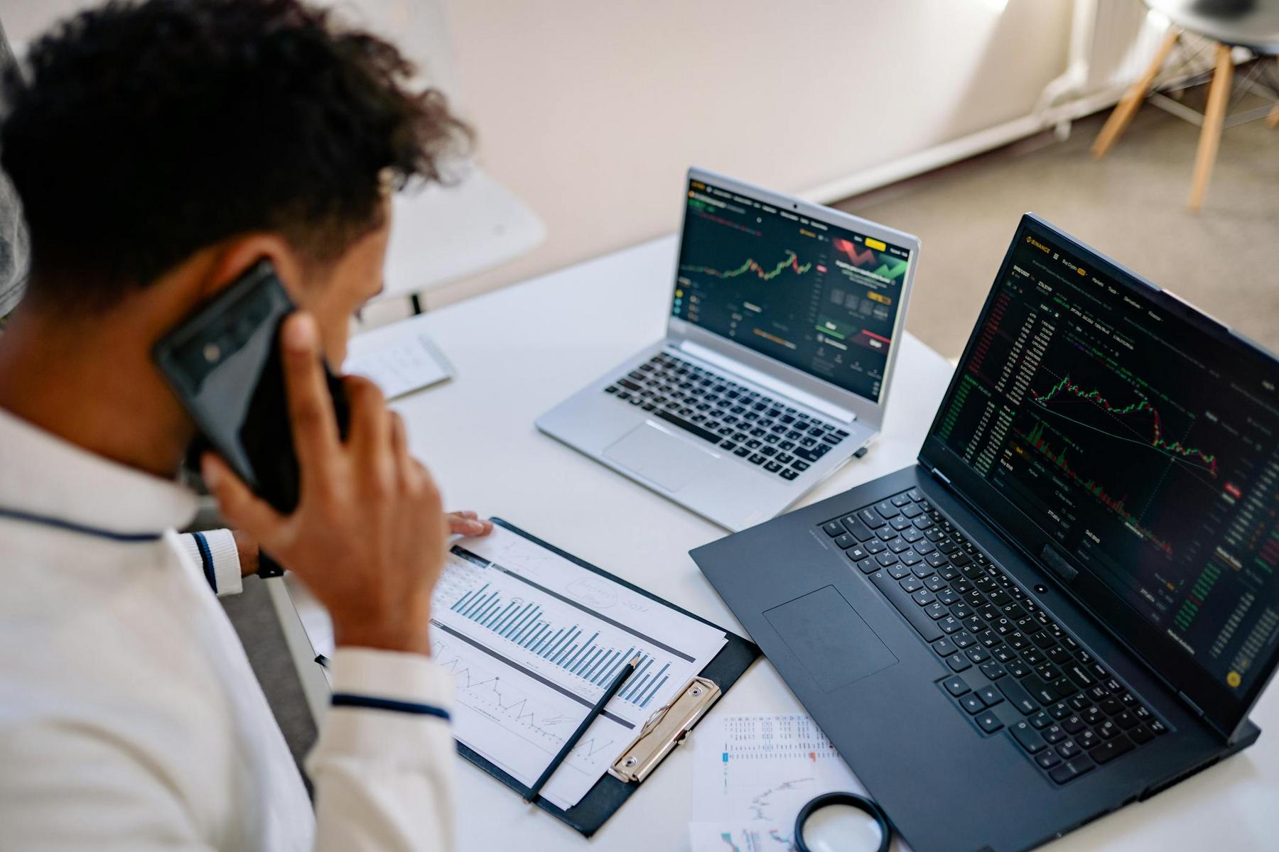A person on the phone reviews printed charts at a desk with two open laptops displaying financial graphs and stock trading data.