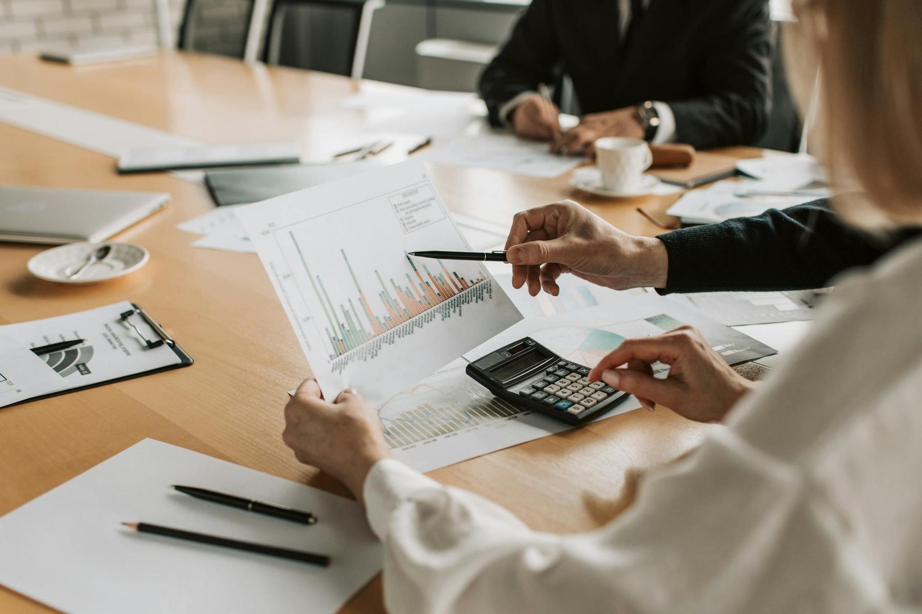 Two people in business attire review a printed bar chart and use a calculator at a conference table covered with documents, pens, and a cup of coffee.