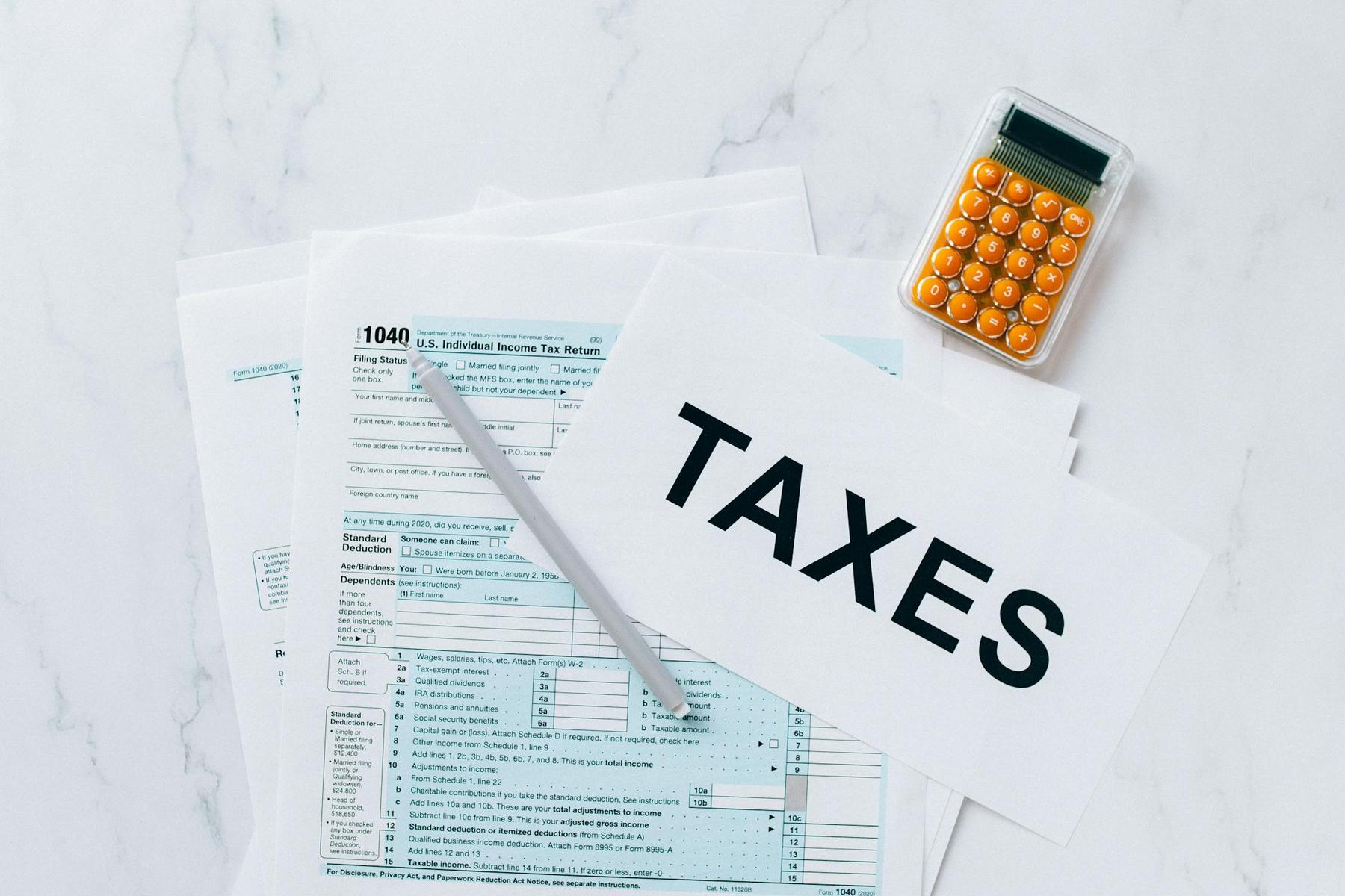 A stack of U.S. tax forms with a pen, a small calculator, and a sheet of paper labeled "TAXES" on a white marble surface.