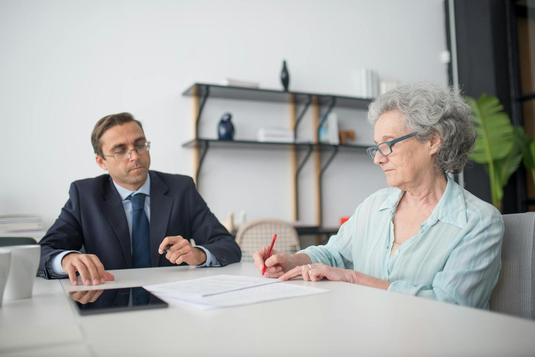 An older woman reviews and signs documents at a desk while a man in a suit sits across from her, observing.